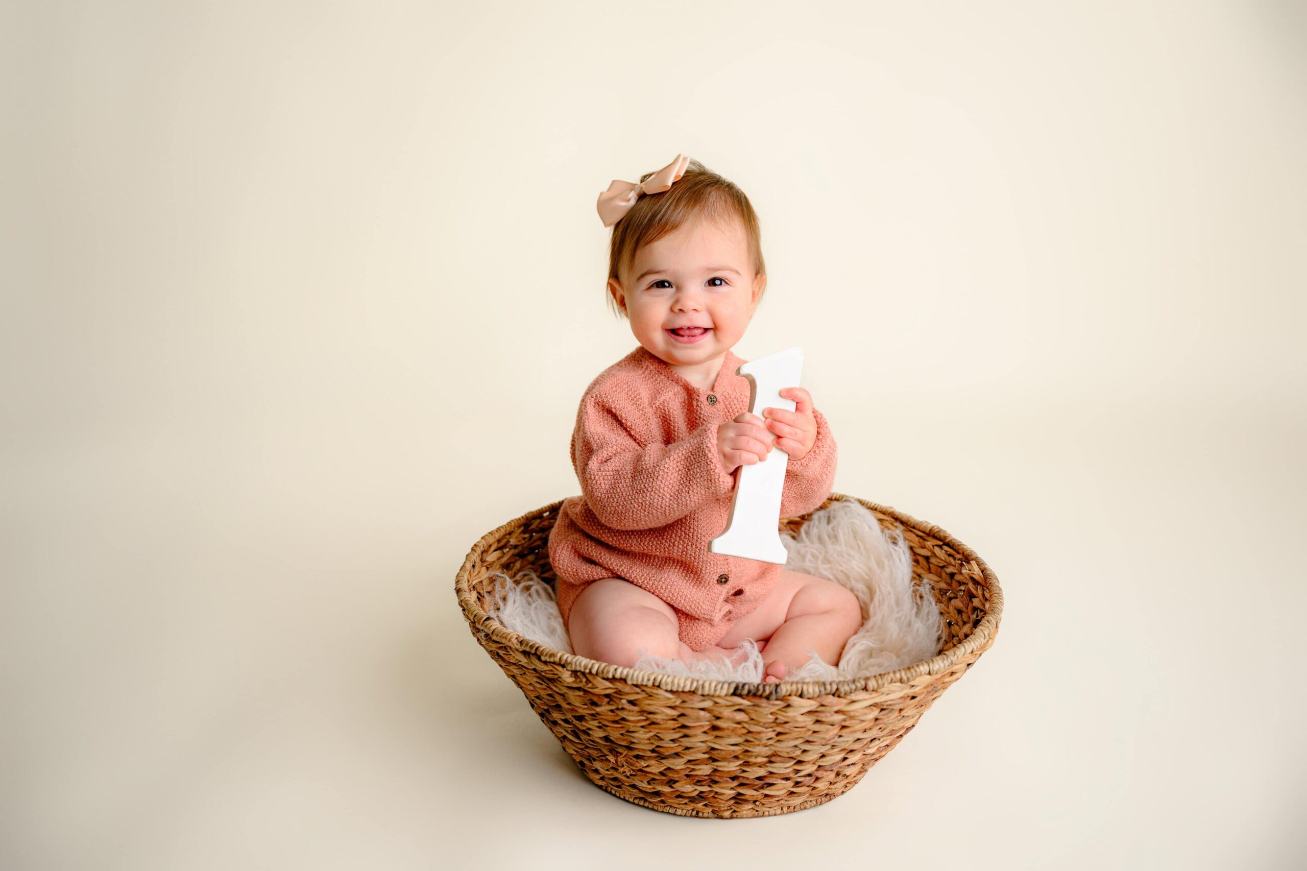 Adorable baby girl sitting in a woven basket, holding a white wooden number '1' and smiling brightly during her simple cake smash photo session