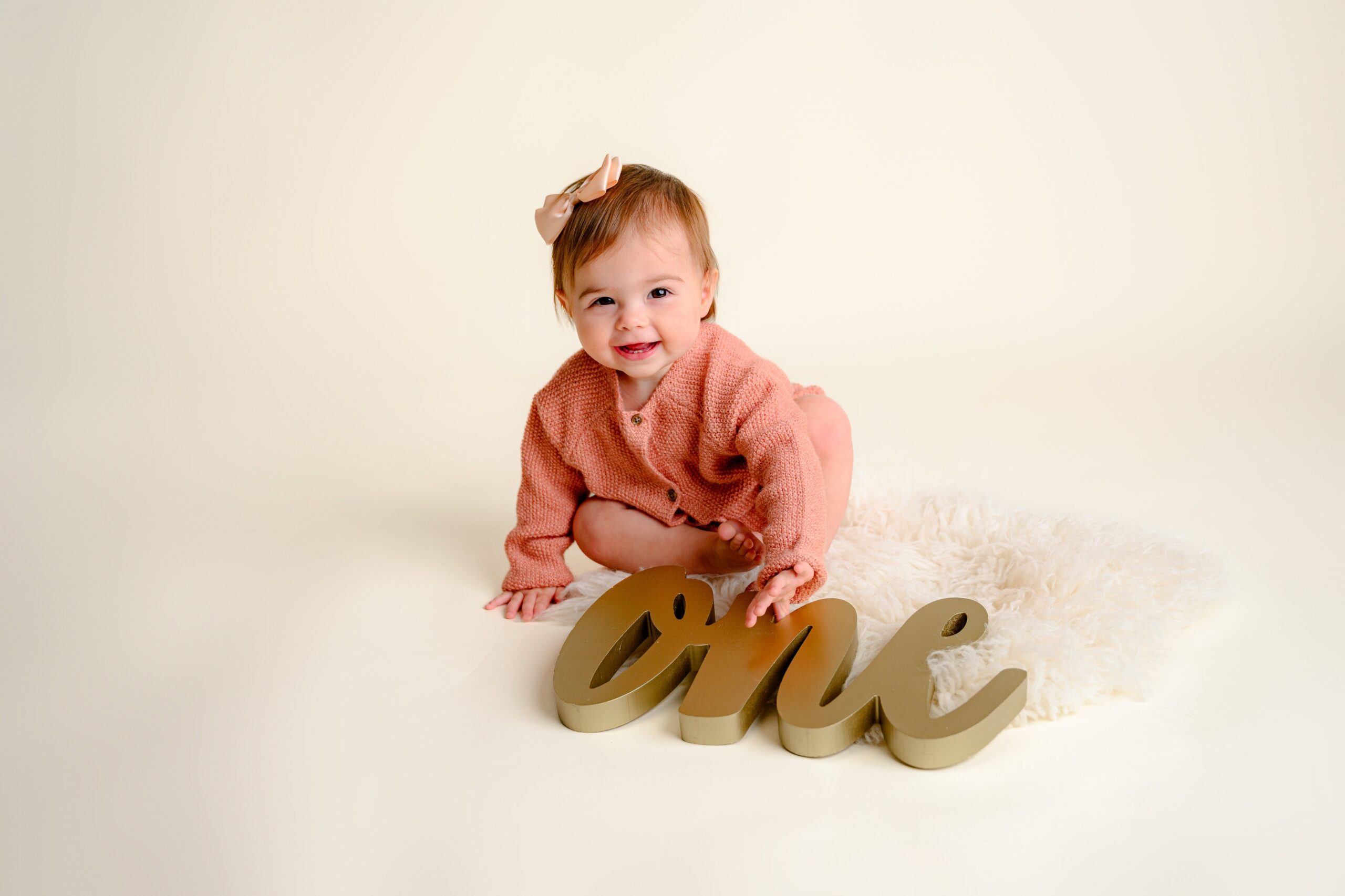 Smiling one-year-old baby girl in a peach sweater, crawling near a gold 'one' sign, surrounded by soft cream-colored décor for her milestone cake smash session