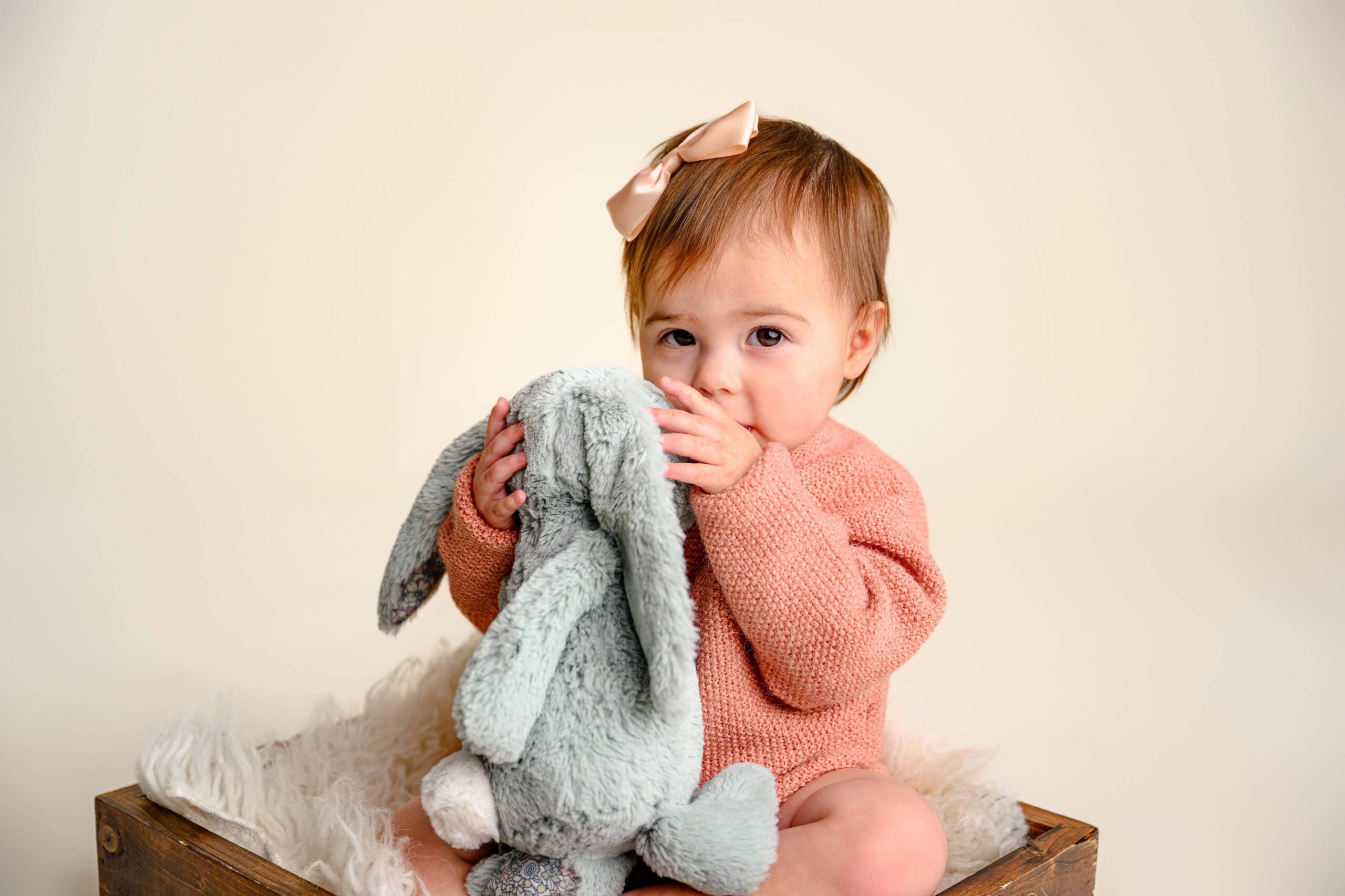 Sweet baby girl hugging a soft gray bunny stuffed toy, wearing a peach knit outfit and a beige bow, captured during her first birthday photoshoot