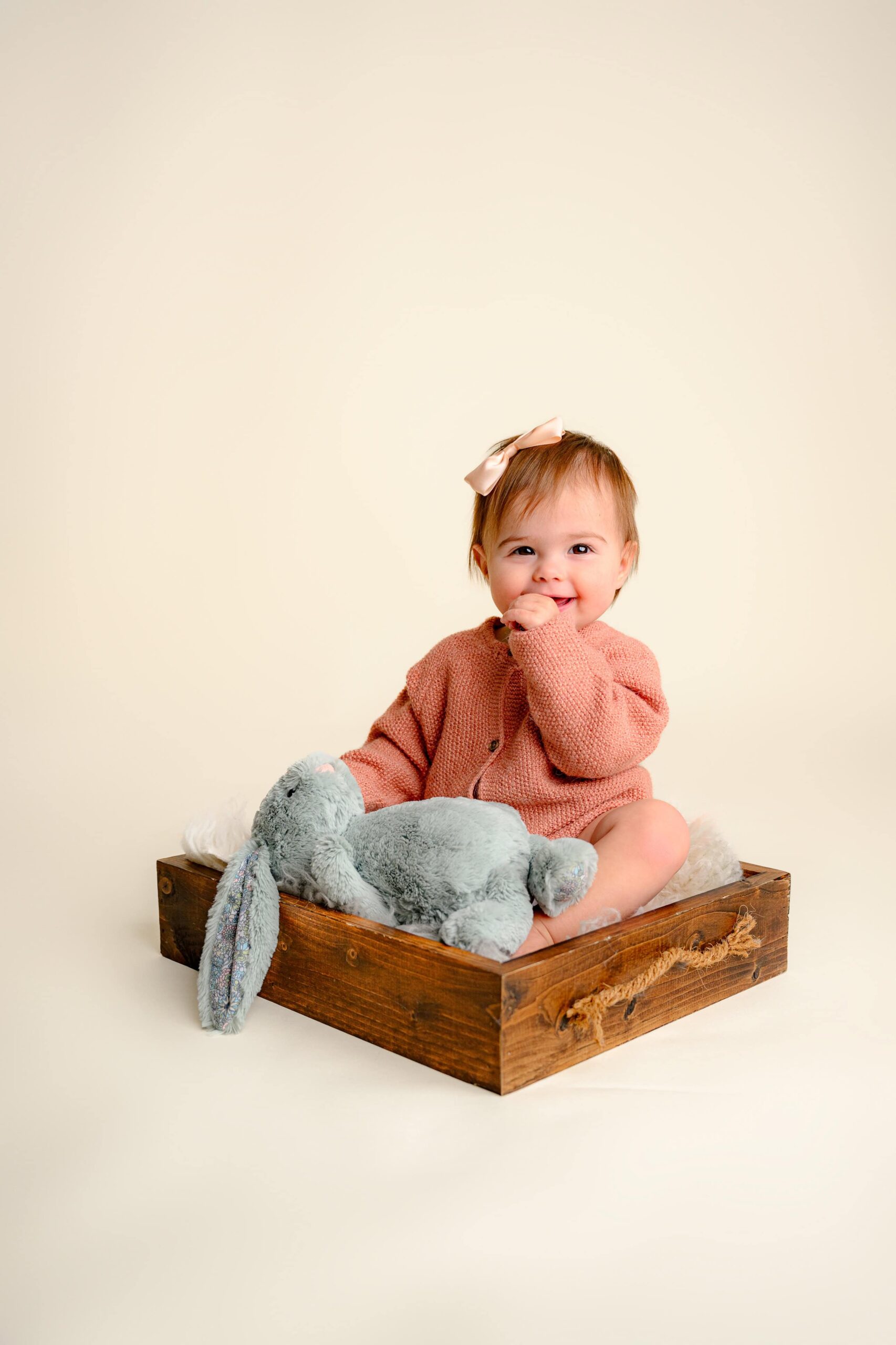 Happy one-year-old baby girl sitting in a rustic wooden crate, holding her gray bunny plush toy, smiling sweetly for her simple cake smash milestone session