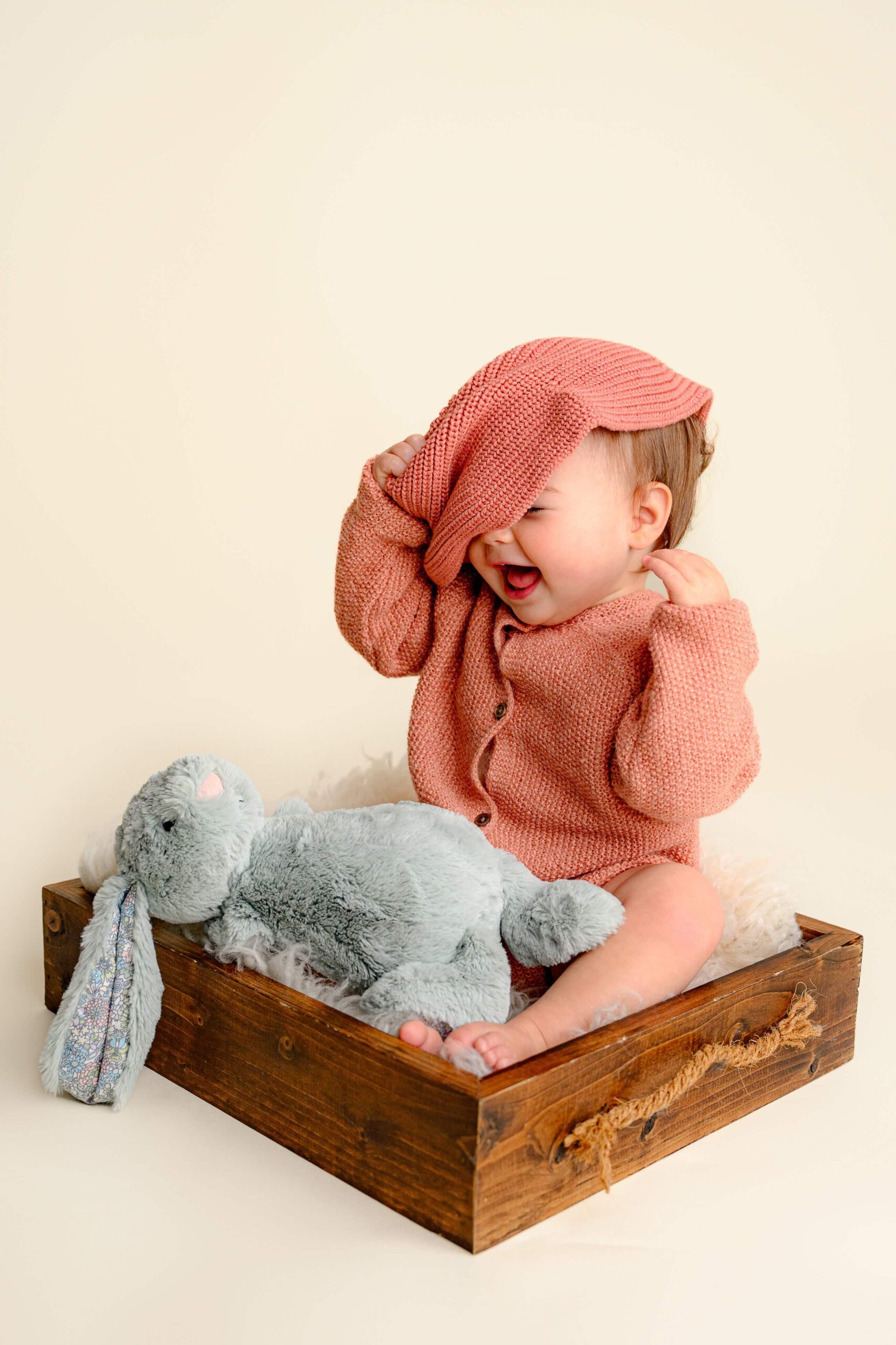 Sweet baby girl sitting with her grey bunny and laughing as she takes off her bonnet, wearing a peach knit outfit and a beige bow, captured during her first birthday photoshoot