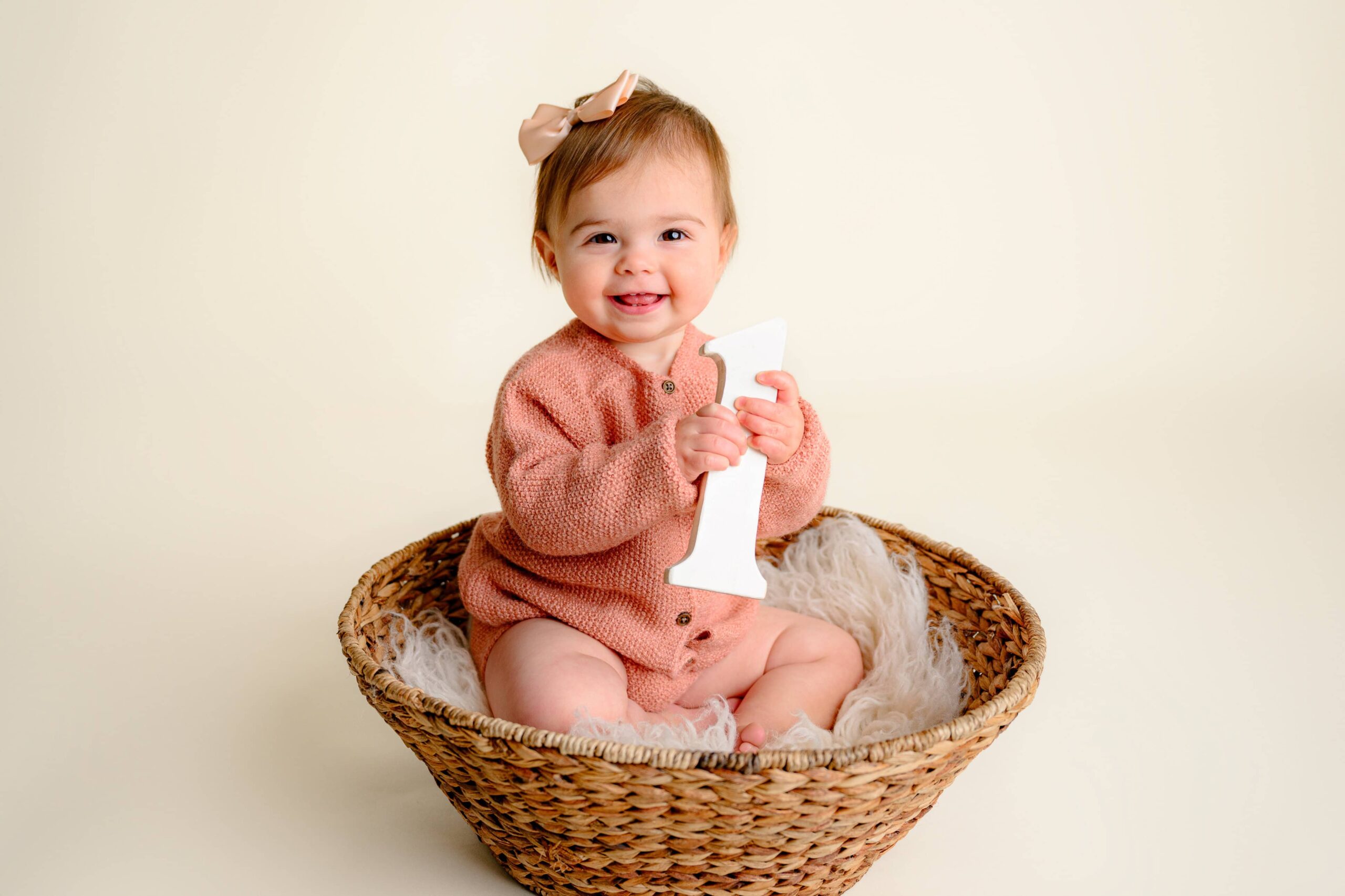 A one year old dressed in a salmon colored sweater and bow in her hair sits in a basket holding a number one prop during her milestone session