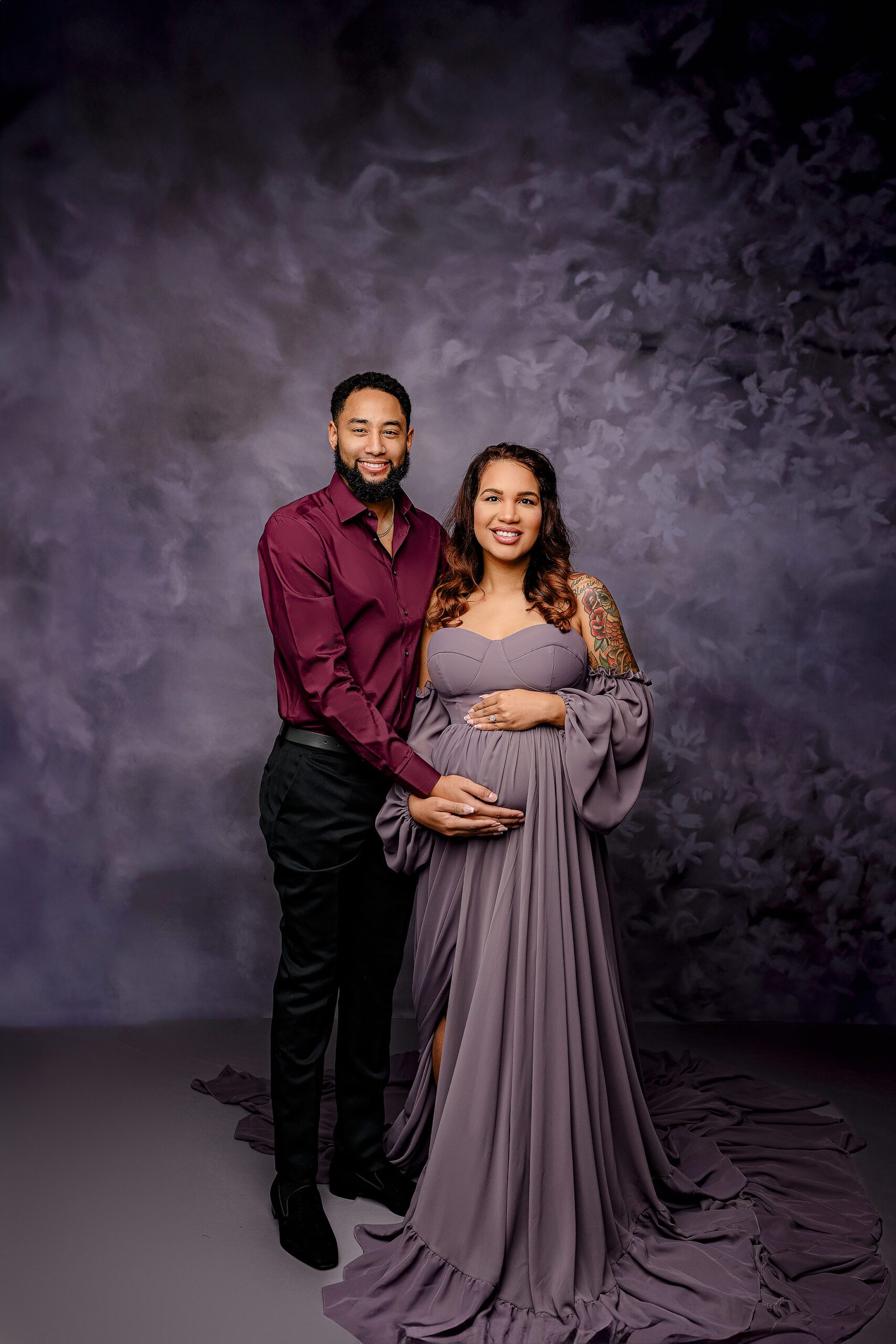 couple stands together in a traditional maternity pose with hands on her belly smiling toward camera with mom wearing a dusty purple dress and dad dressed in maroon on a purple set up