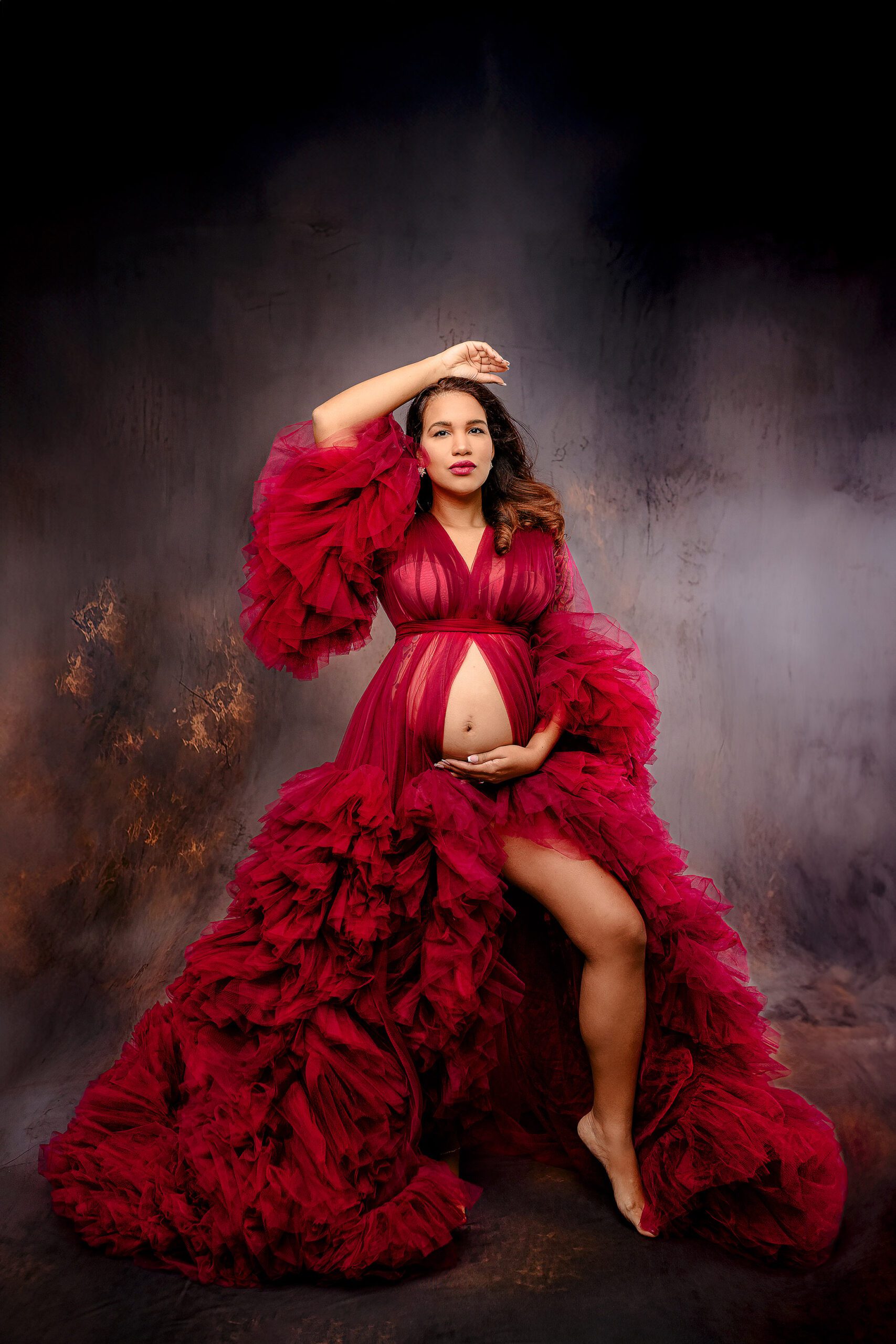 mom-to-be poses dramatically in red tulle gown during her studio maternity studio session with one leg out and her hand above her head