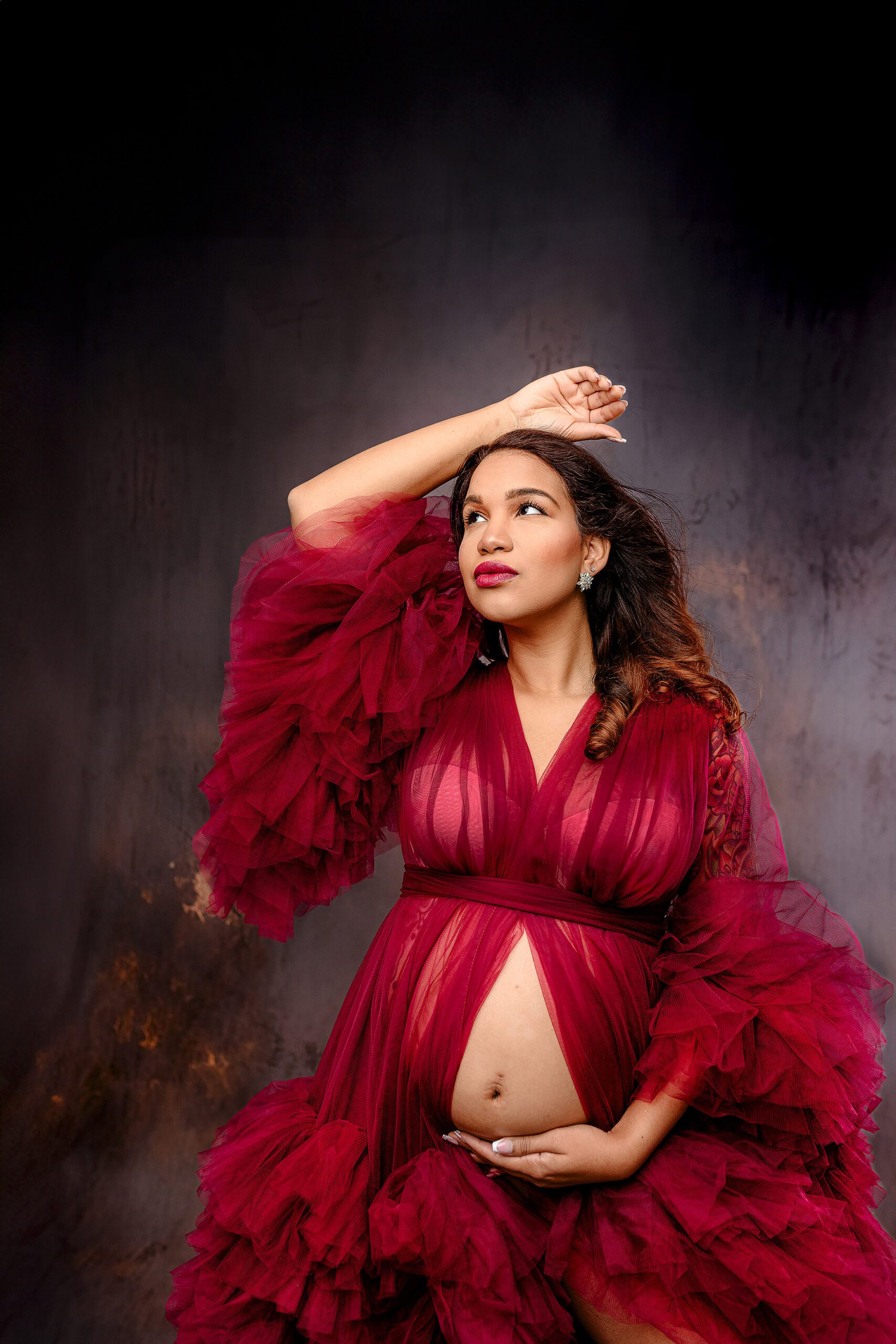 mom-to-be poses dramatically in red tulle gown during her studio maternity studio session with her hand above her head