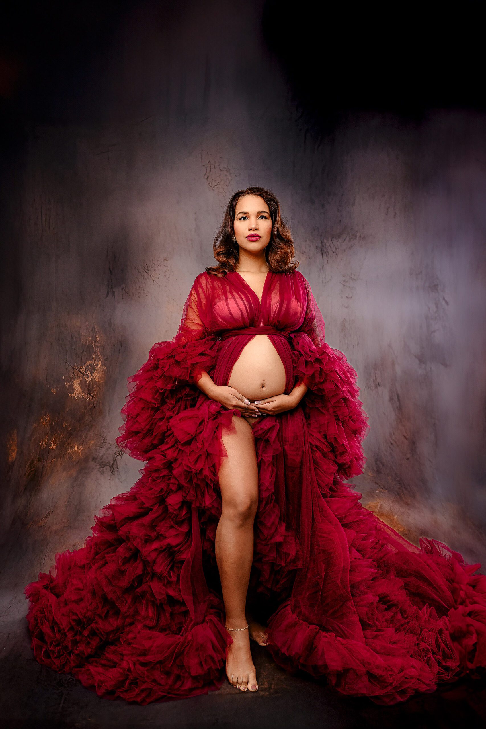 charlotte, nc mom-to-be poses dramatically in red tulle gown during her studio maternity studio session with her hands on her belly and her belly exposed and leg out