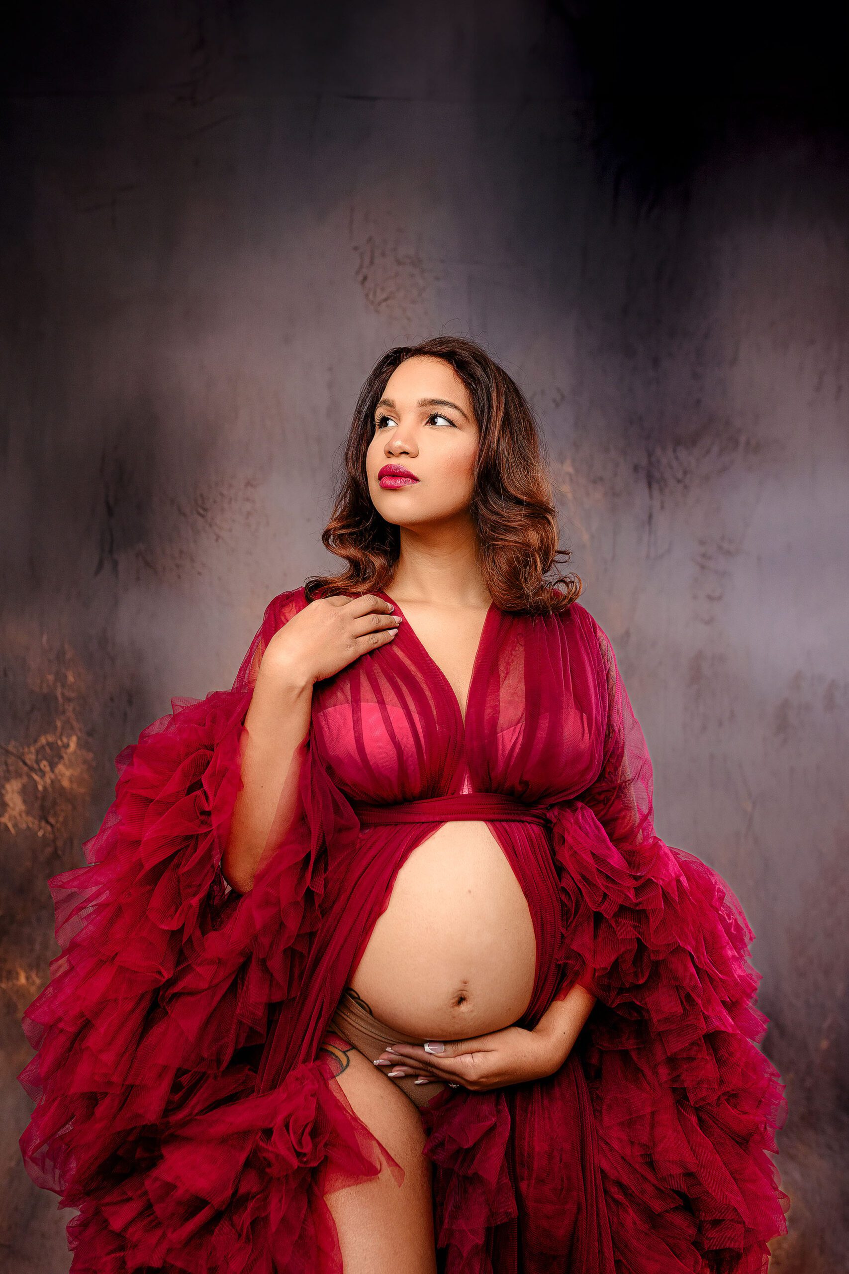 mom-to-be poses dramatically in red tulle gown during her studio maternity studio session with her belly showing and her hand on her collarbone and her belly