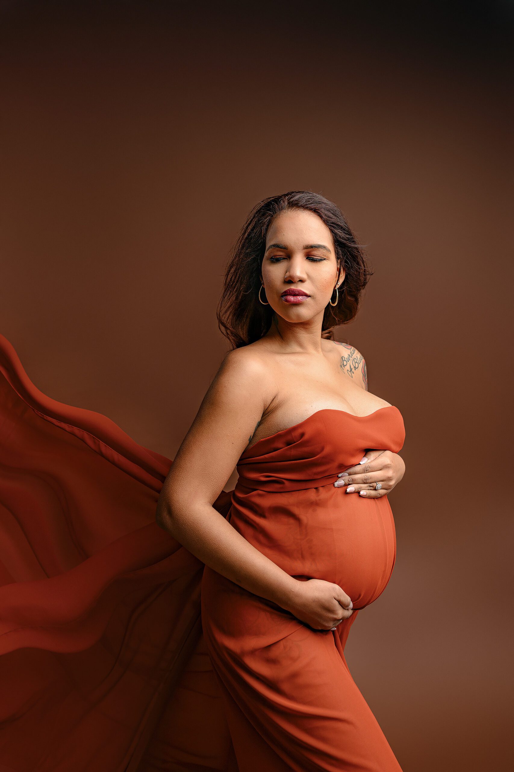 Ballantyne mom-to-be is wrapped in a rust chiffon scarf posed looking toward her shoulder  fabric flowing behind her in the waxhaw nc studio