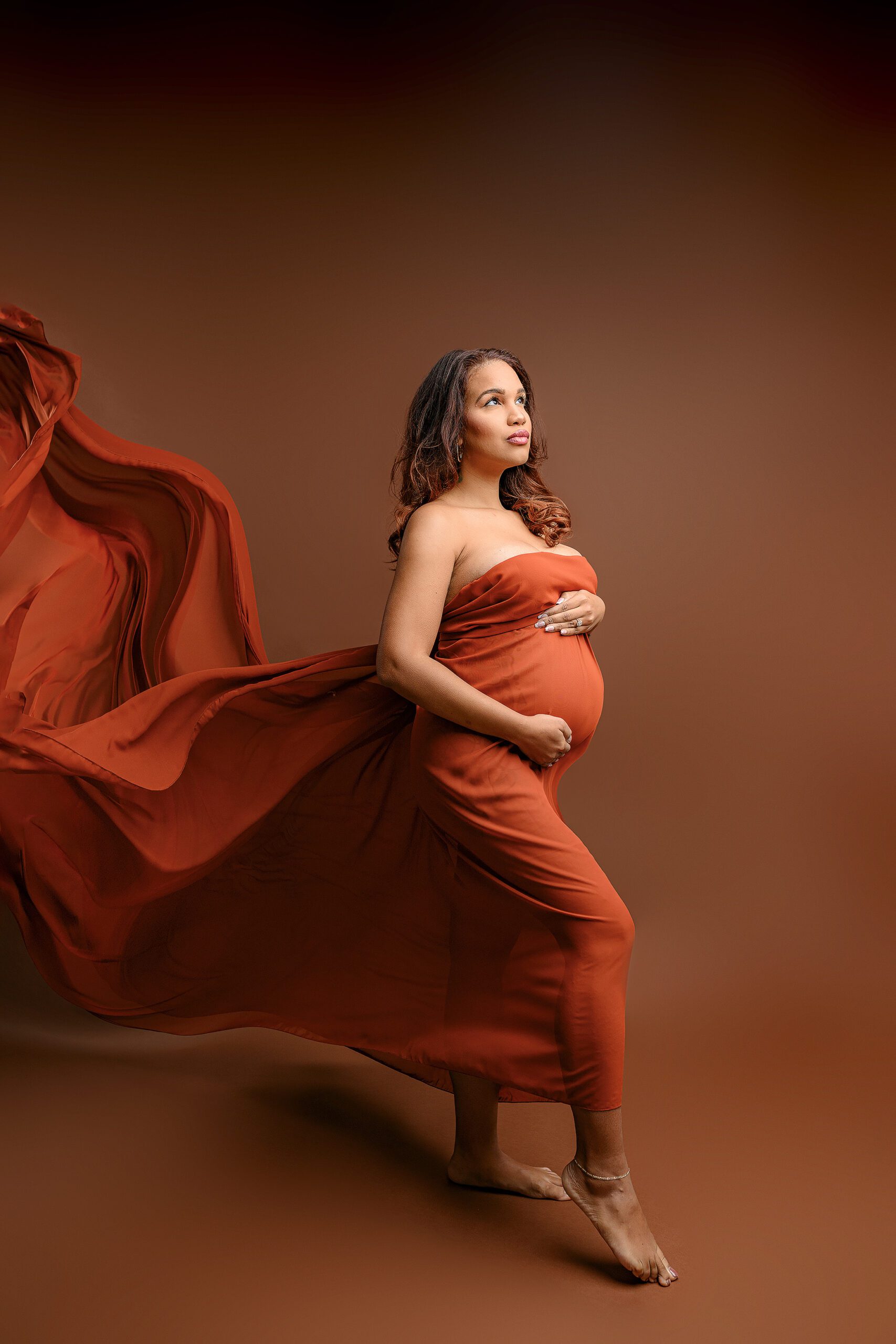 Ballantyne mom-to-be is wrapped in a rust chiffon scarf posed looking toward the light with the fabric flowing behind her in the waxhaw nc studio