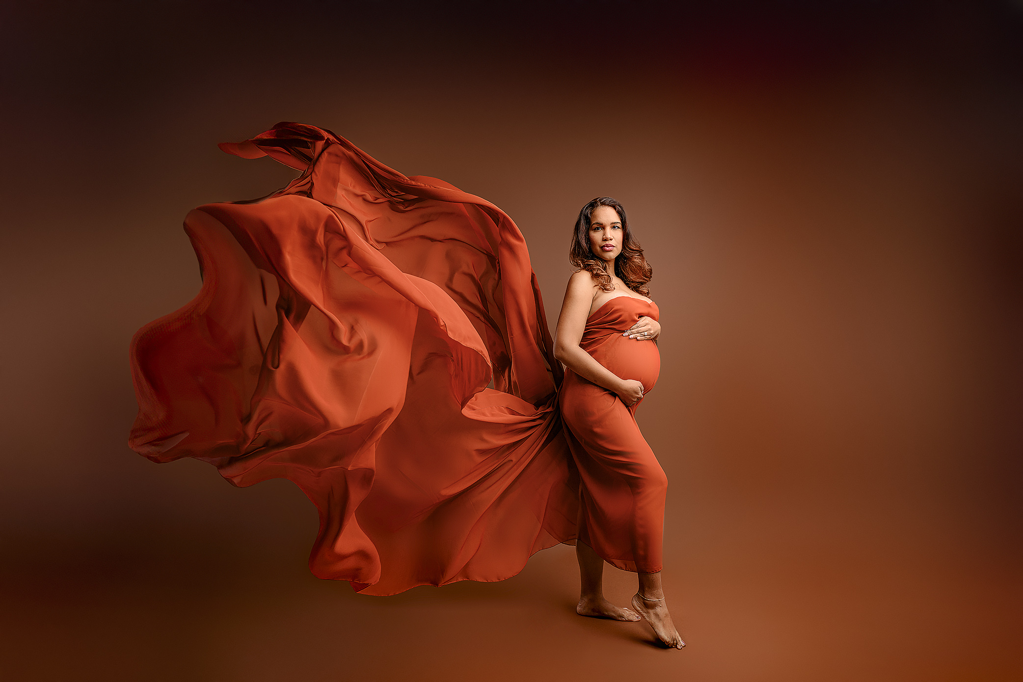 Mom-to-be Monet Watson standing posed wrapped in a rust colored chiffon wrap flowing behind her in the waxhaw, nc studio on a brown background