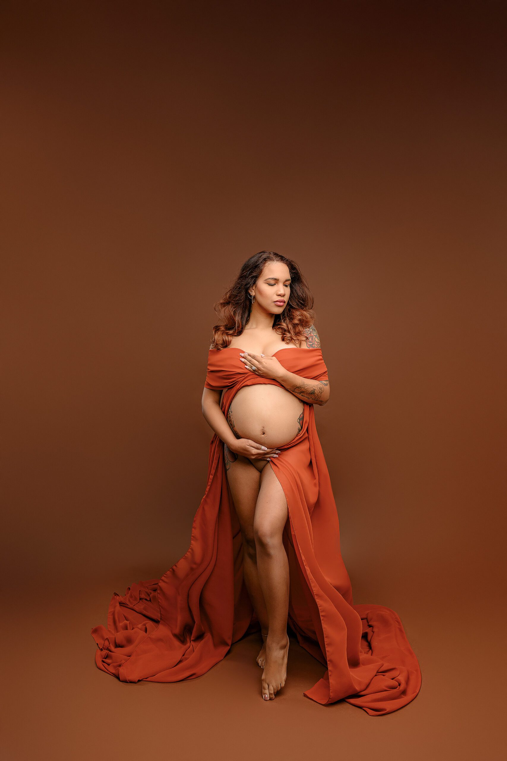 Ballantyne mom-to-be is wrapped in a rust chiffon scarf posed looking down toward her shoulder  with her belly exposed and wrap over her shoulders