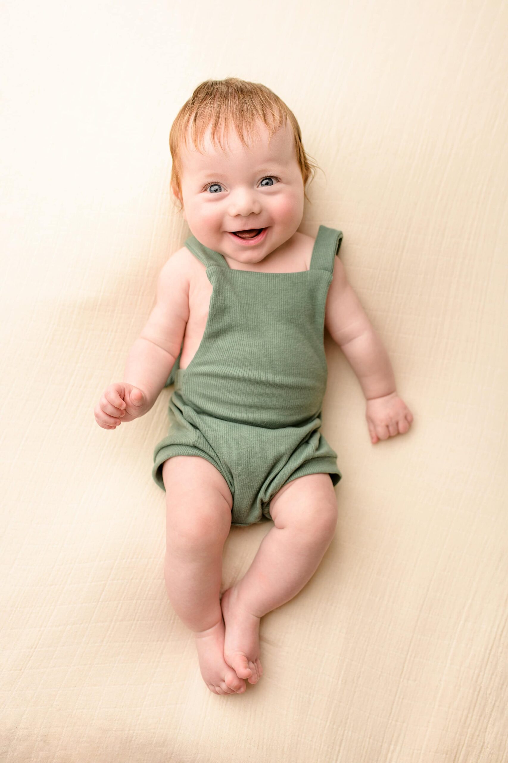 You can almost hear the giggle of this little redheaded boy with bright blue eyes laying on a neutral background looking up with the camera while wearing a green bubble romper