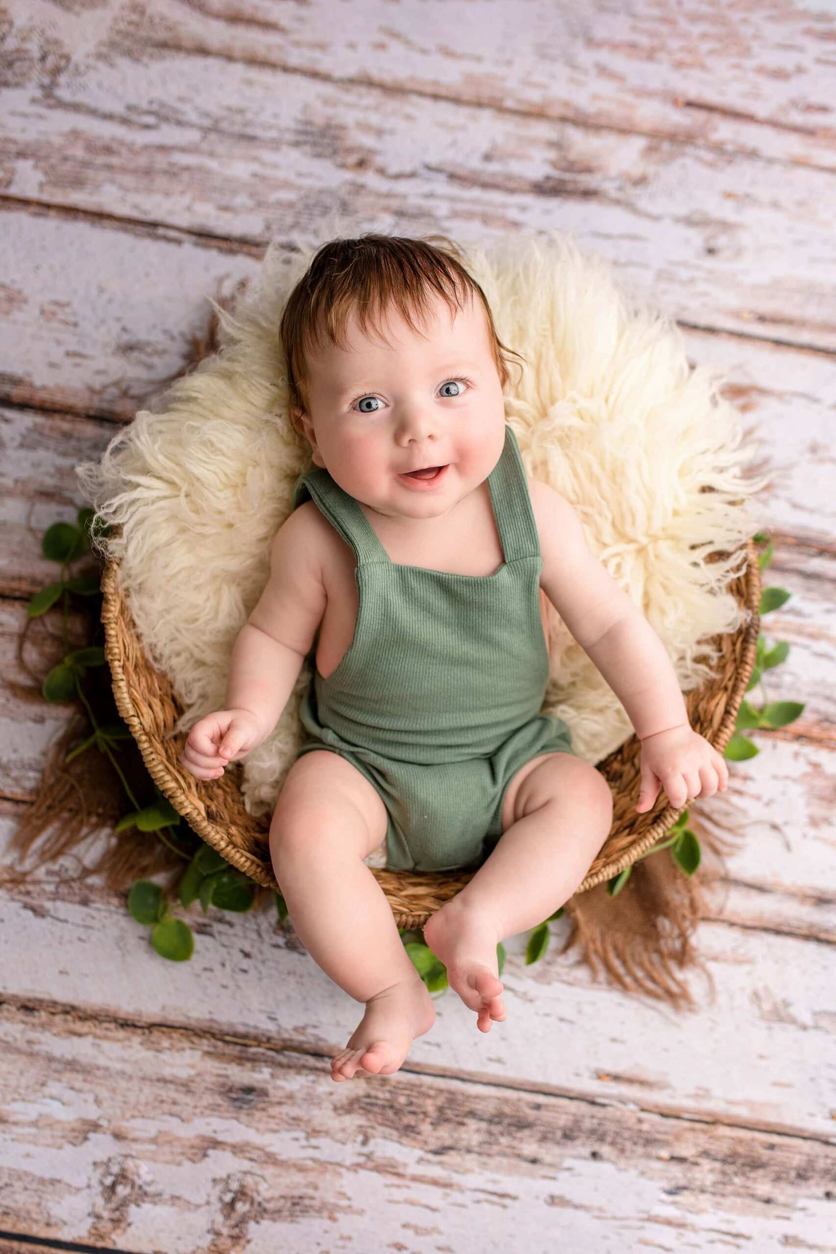Little boy smiles he fits in a basket wearing a sage green bubble overalls during his baby photos