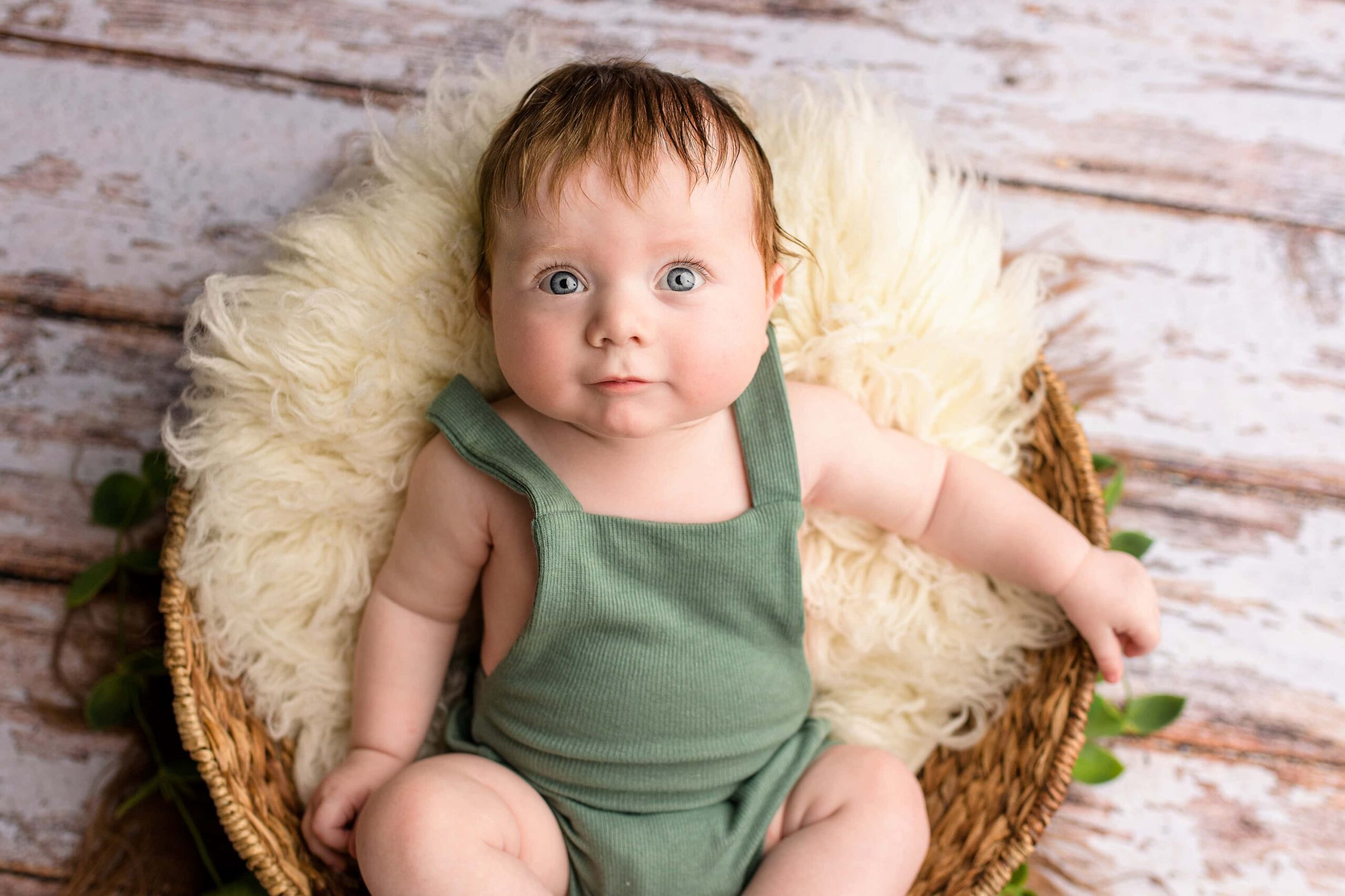 Dressed in a green bubble romper this four month old redhead boy with blue eyes is placed in a basket with neutral colored fur to celebrate his milestone