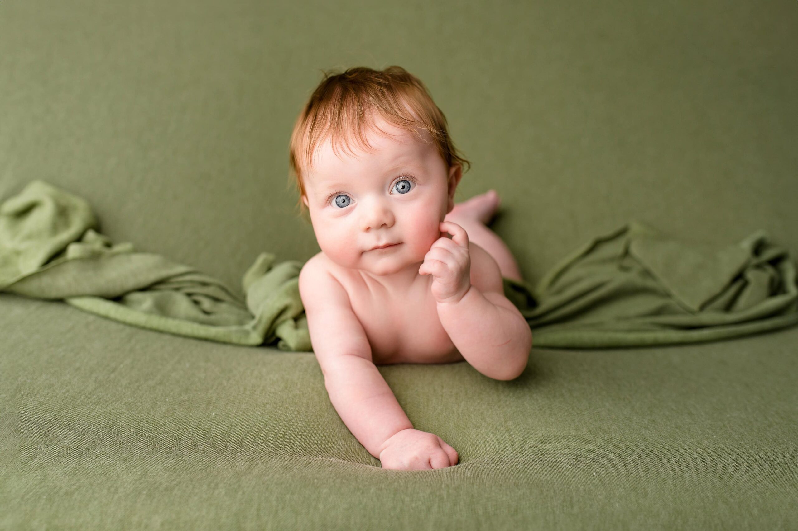 Little redheaded boy is surrounded in a sage green background which compliments his skin tone while laying on his belly showing off his new four month old skills of pushing up and smiling
