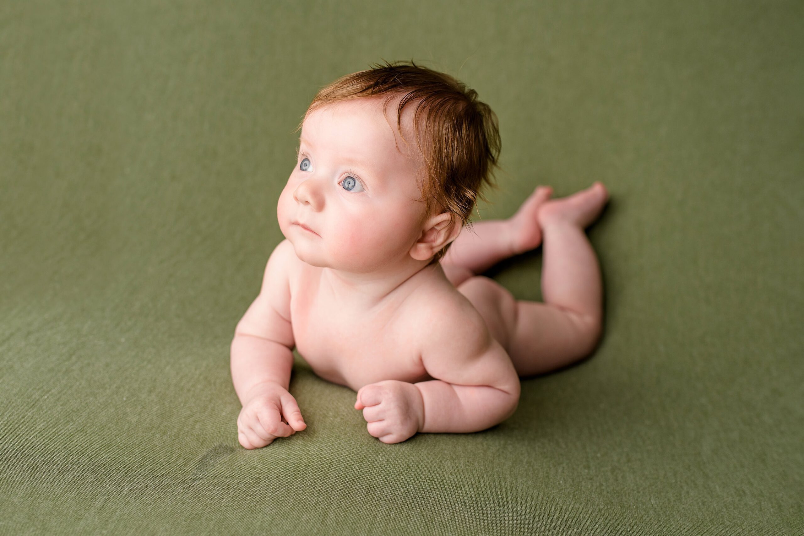 Sweet little photos of a four month old baby boy looking off camera while laying on his belly on a green background