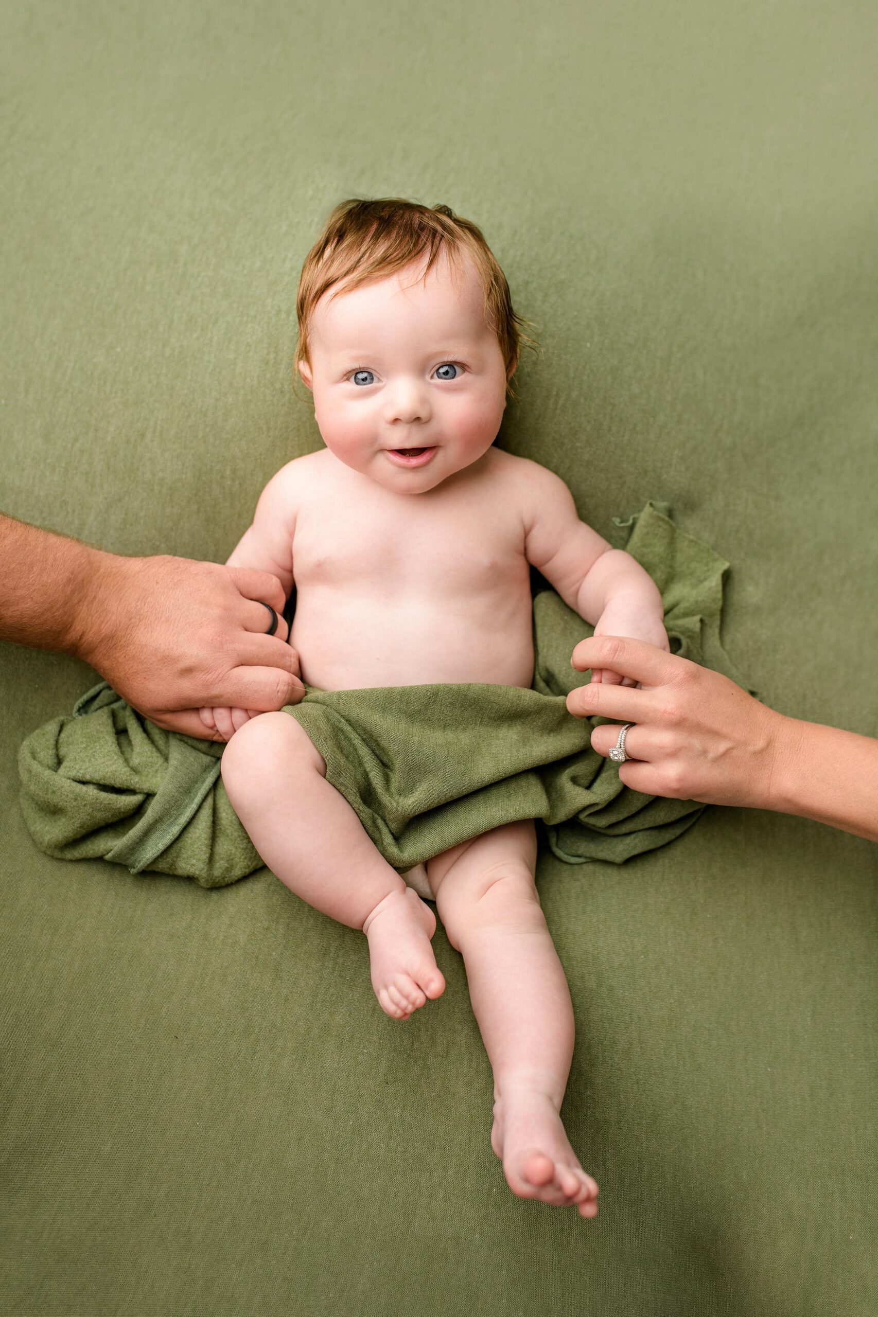 Holding his mom and dad‘s hands, the little boy lays gently on a green background during his baby photos