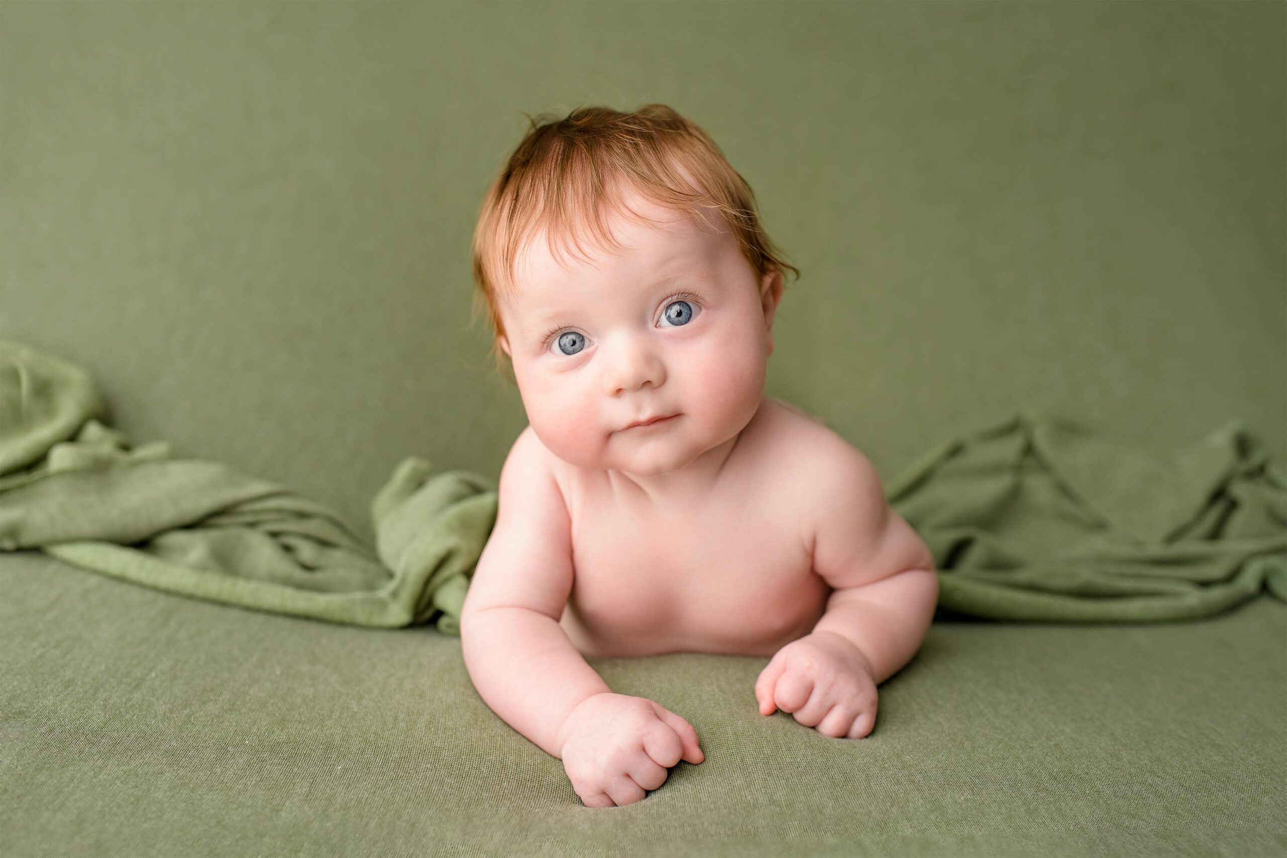 A sweet image of a four month old redheaded boy with blue eyes posed gently on his belly on a green background