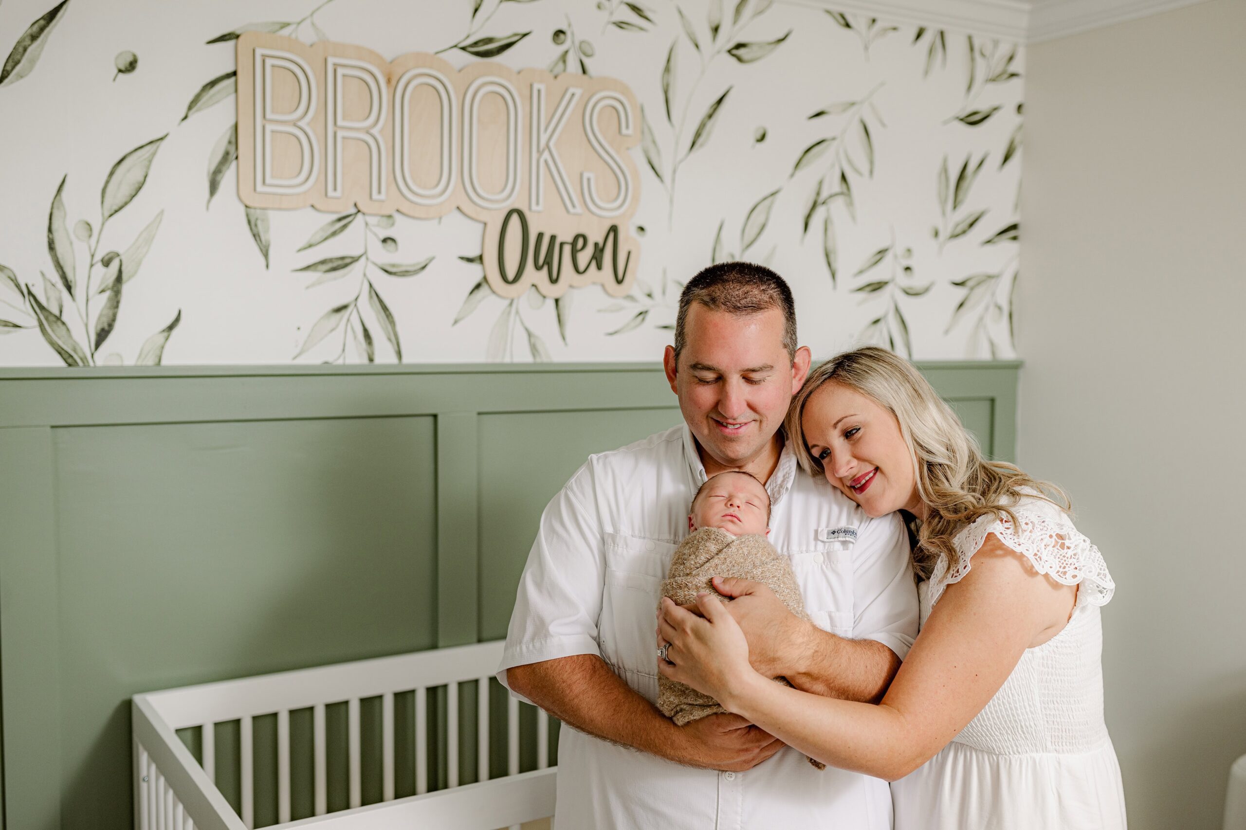 dad holds baby boy in his arms while the newborn is wrapped in a neutral wrap and mom lays her head on dad's shoulder in their little boy's nursery with the baby's name on the wall behind them