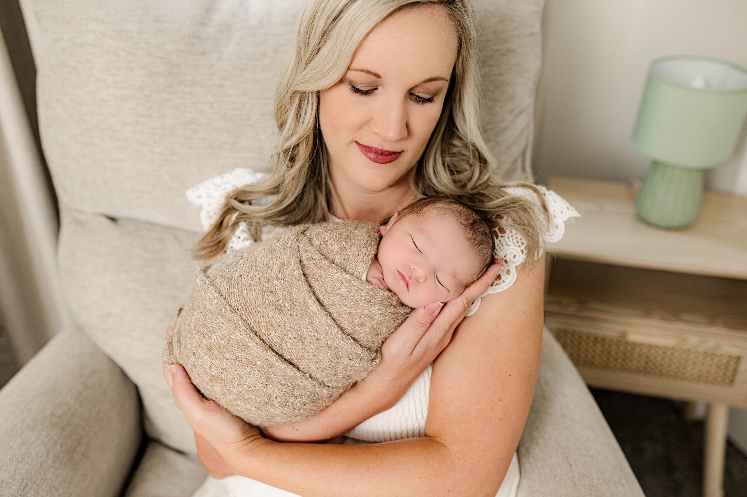 blonde hair mom dressed in cream sits in chair cradling her newborn boy named Brooks. Brooks is wrapped in a brown wrap to represent his nursery and the natural tones requested by mom for the lifestyle newborn session