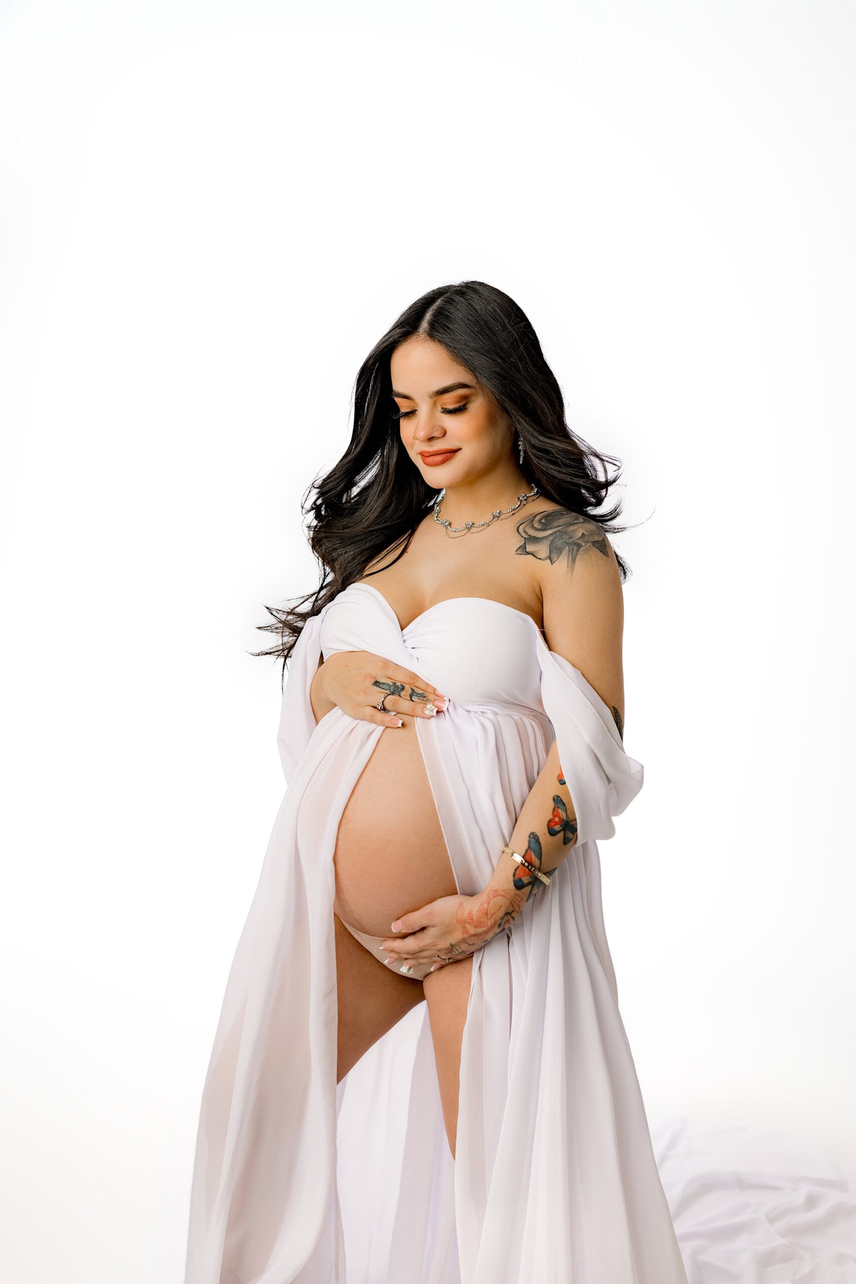 A dramatic portrait of a dark-haired mom-to-be wearing a flowing white dress with a draped sleeve. The dress is gently opened, showcasing her silhouette and her baby bump as she looks down smiling at her growing belly.