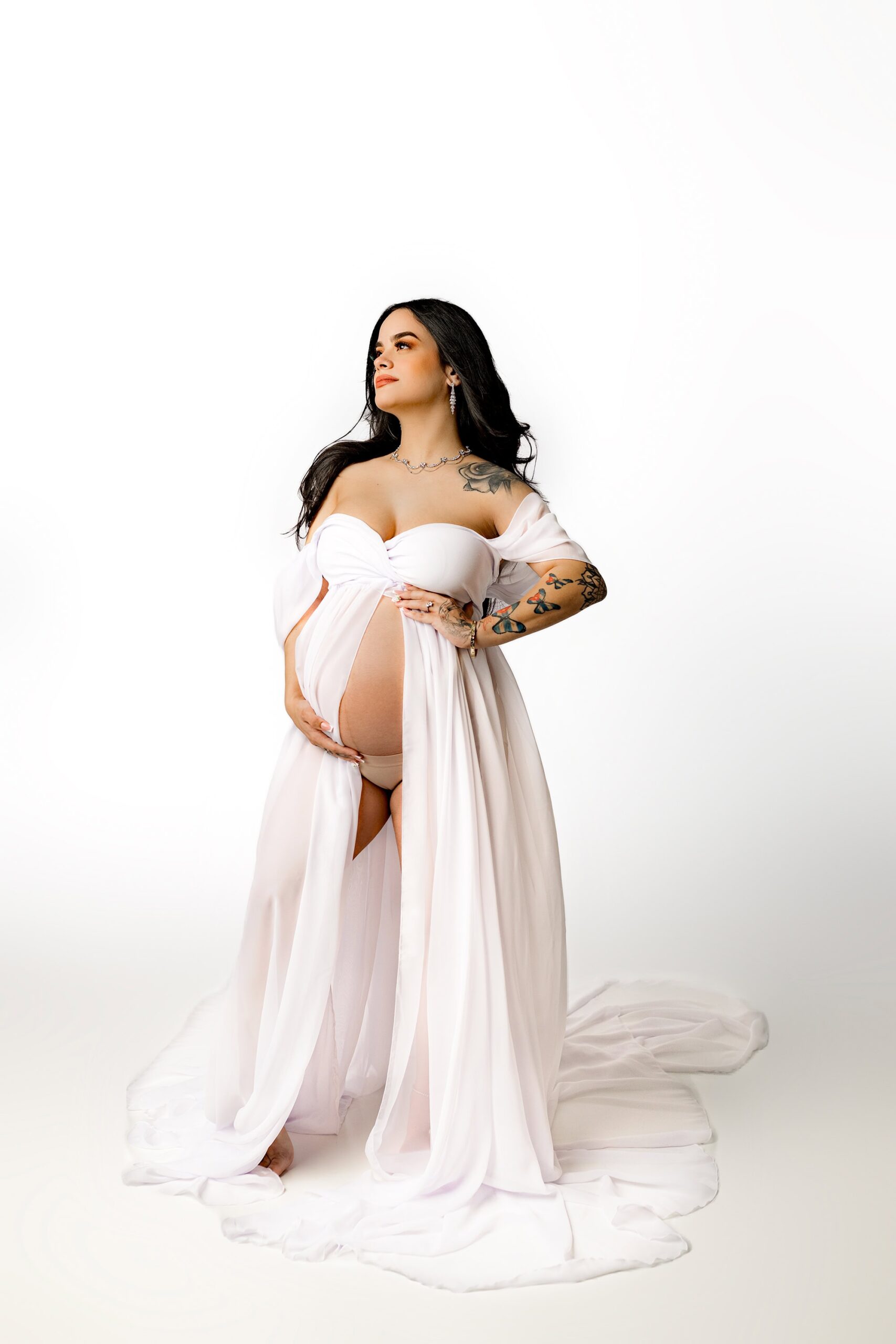 striking image of a dark-haired Charlotte, NC Area Mom-to-Be , dressed in a white gown with a draped sleeve, gently parted to reveal a graceful silhouette. With her hands resting on her face and her gaze directed at the camera, she exudes a sense of calm strength. Her flowing hair adds to the beauty of the moment, creating a timeless feel in this intimate maternity portrait.