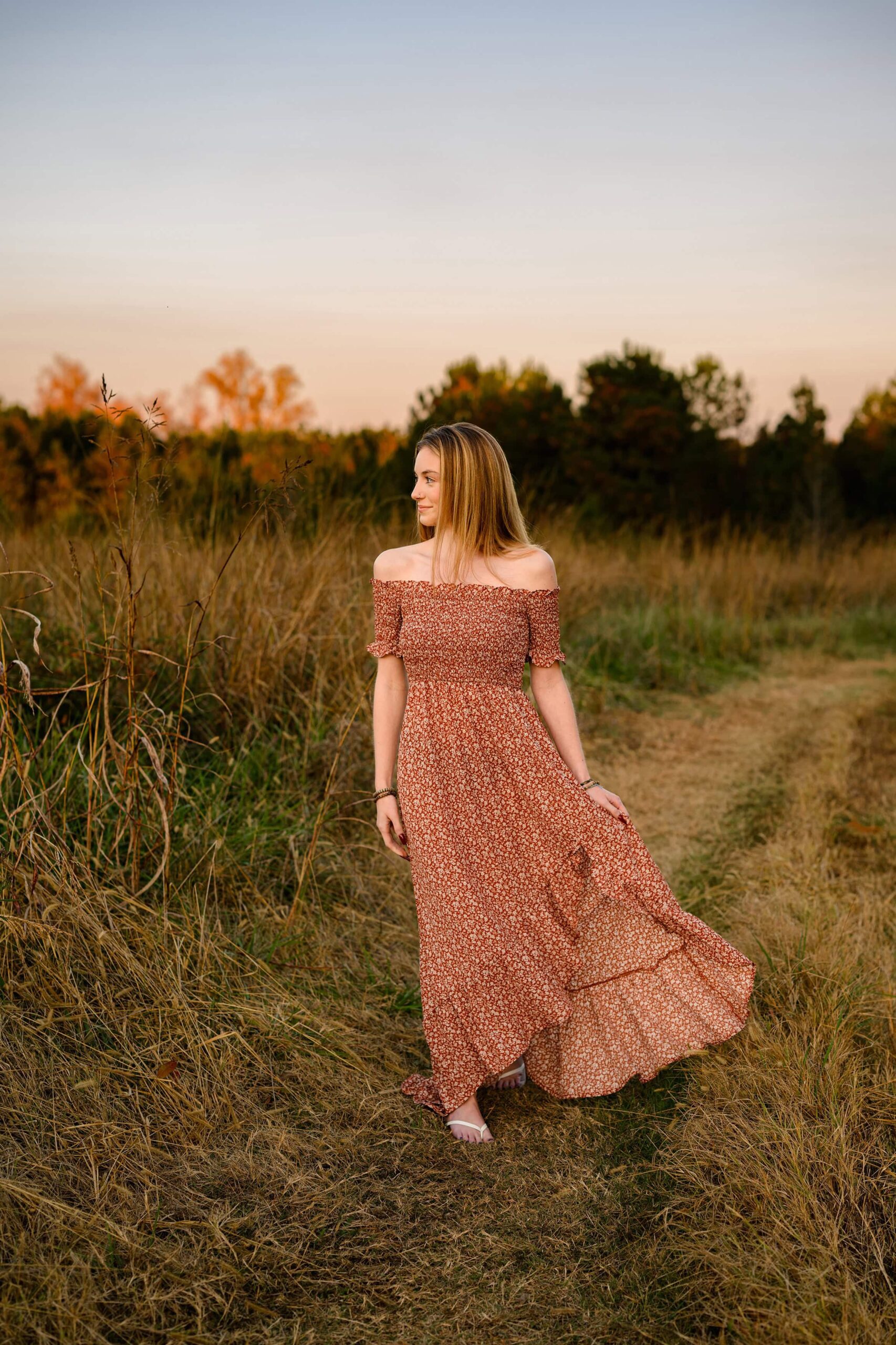senior girl walks down path looking out in a field wearing a rust dress that is flowing in the wind as she walks