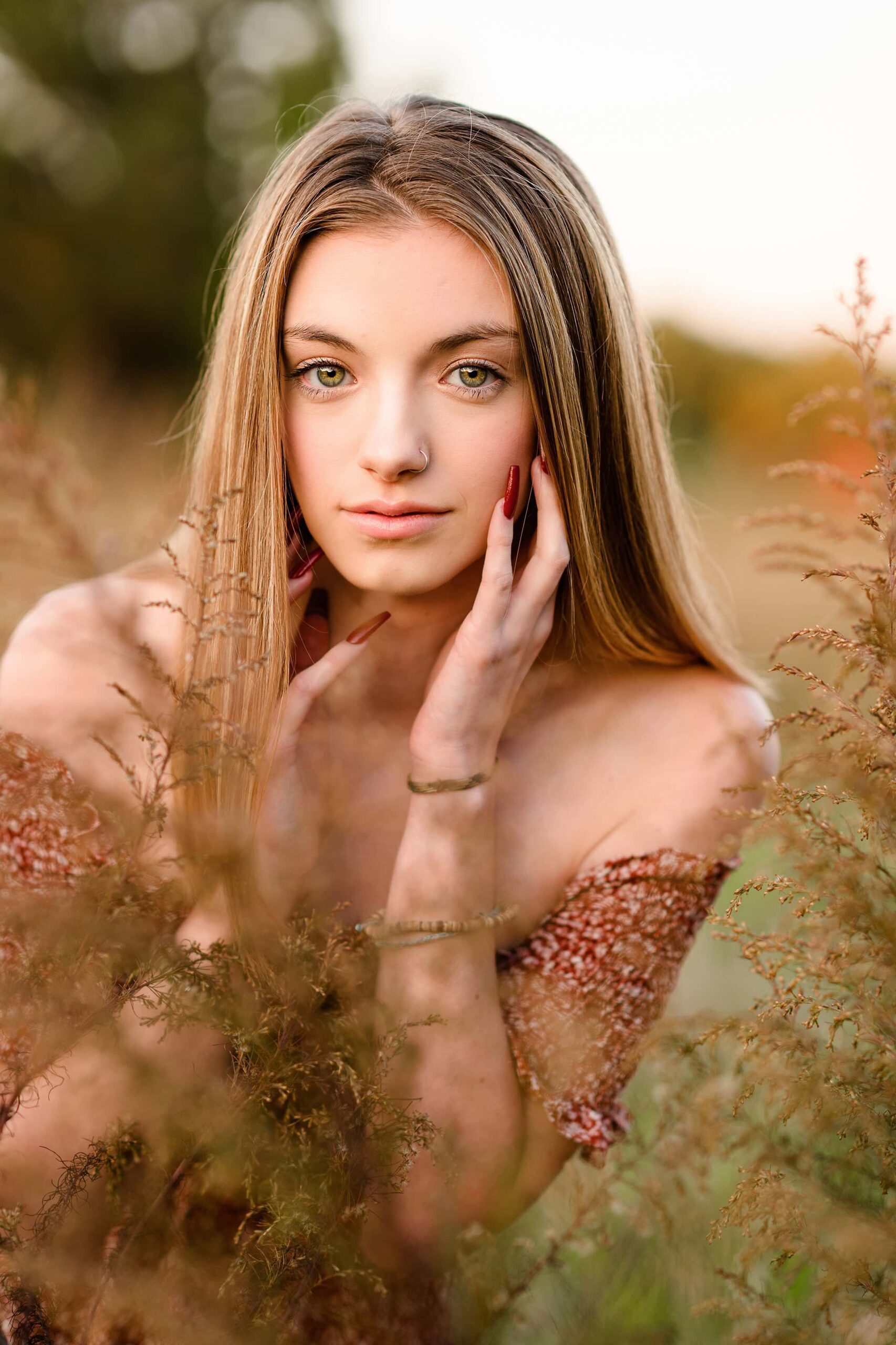 blonde long hair senior girl has hands up by her face wearing a rust colored dress with tall field grasses around her