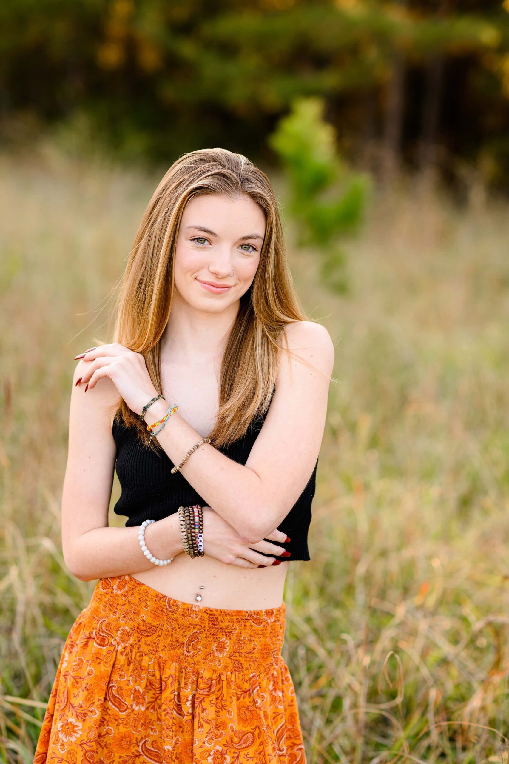 girl grad crosses arm over her black crop top and orange skirt in a field