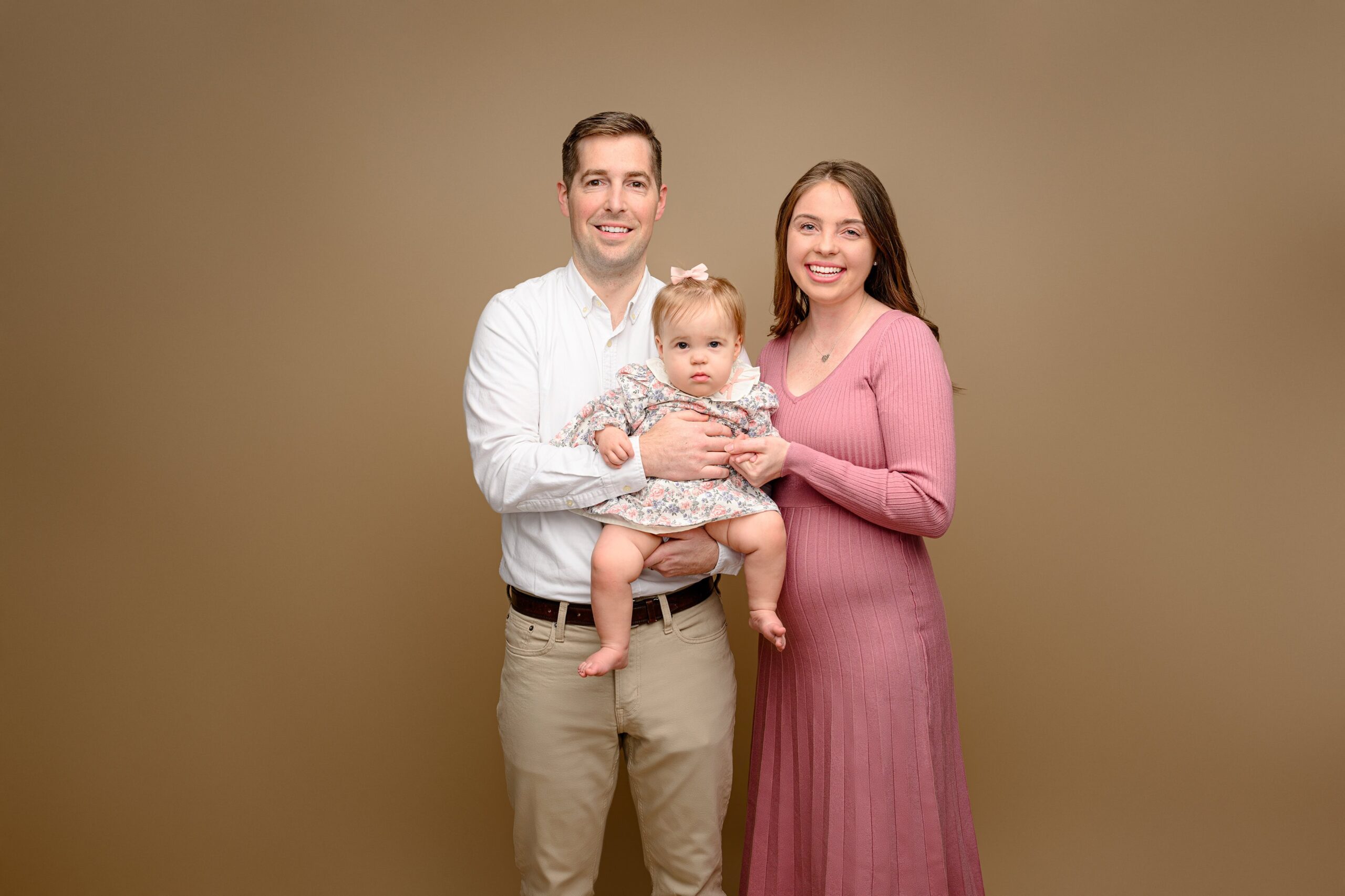 photo including a family of three where dad is classically dressed in khaki pants and a white shirt. mom wears a pink dress showing her new to arrive baby bump. their one year old daughter is held between then wearing a flower dress