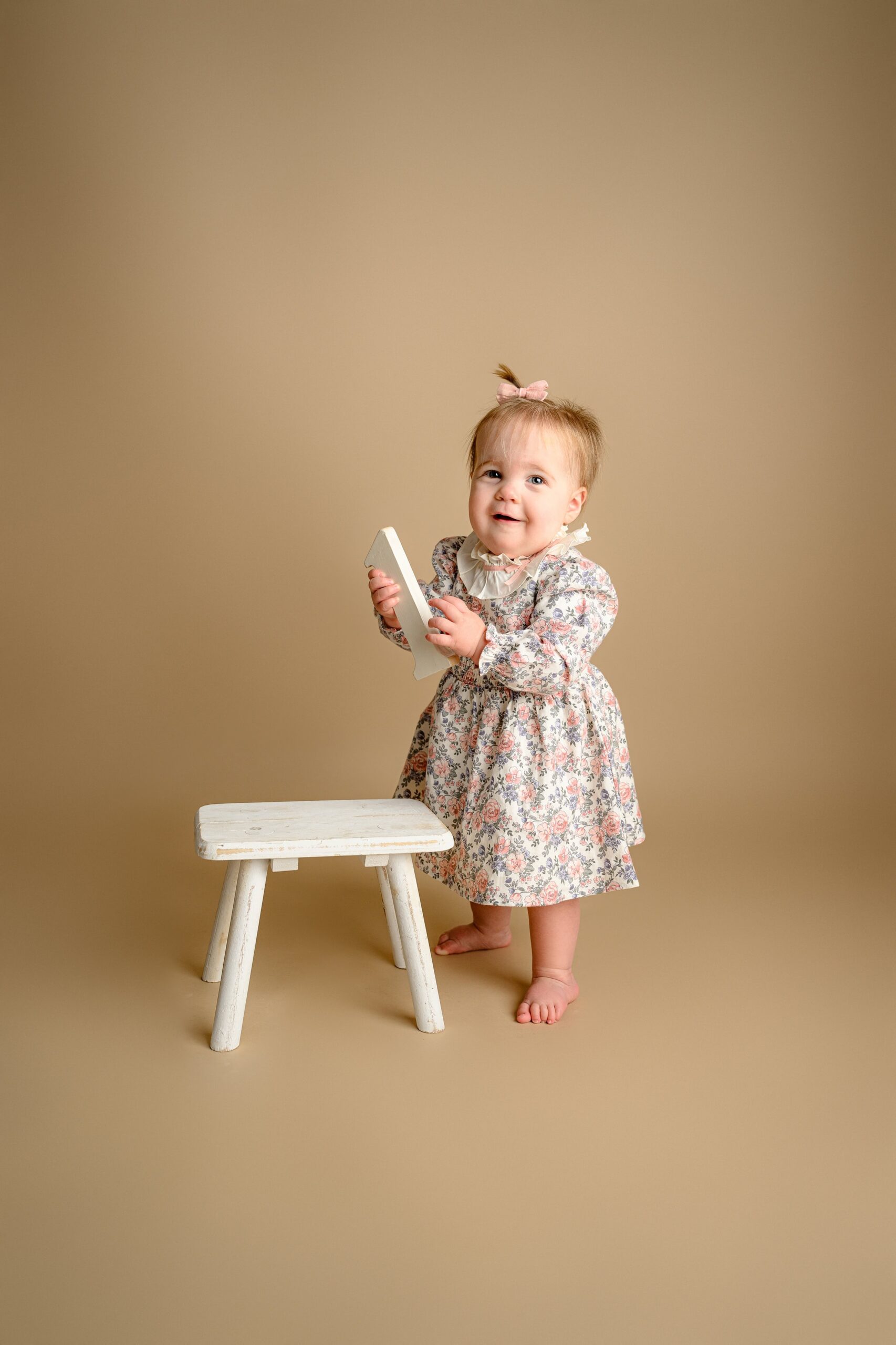 little girl standing solo in a flower dress with her hair pulled back in a little pink bow and holding a number one prop