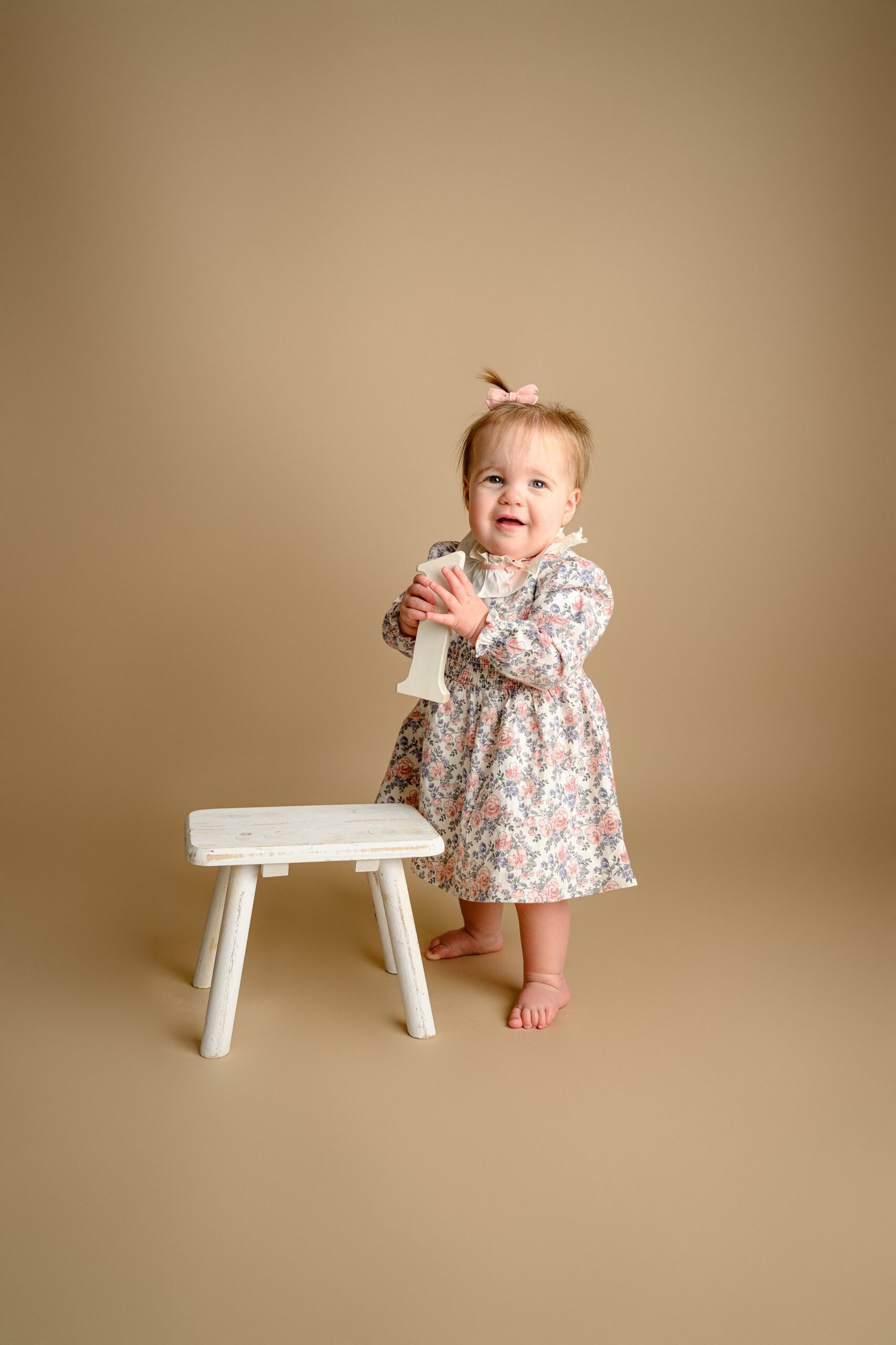 little girl proudly stands next to a stool wearing a flower dresss with her hair pulled back in a little pink bow and holding a number one prop with both hands