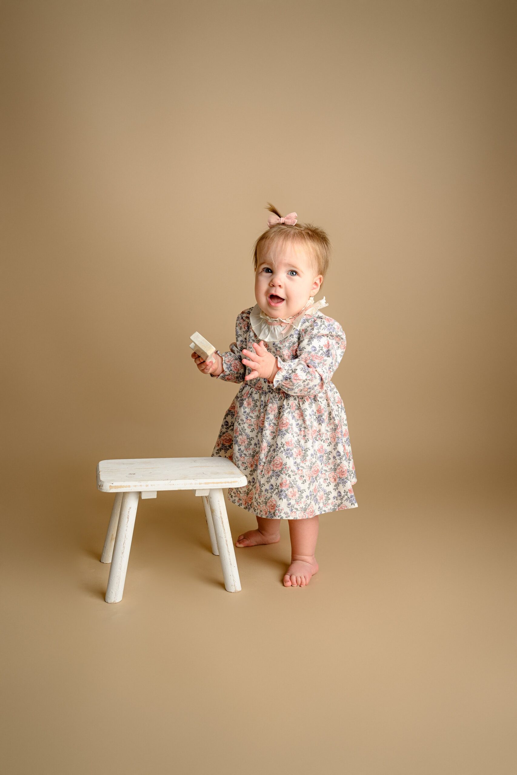 a sweet surprise face on a one year old girl wearing a flower dress as she stands for the first time without assistance during her baby photos