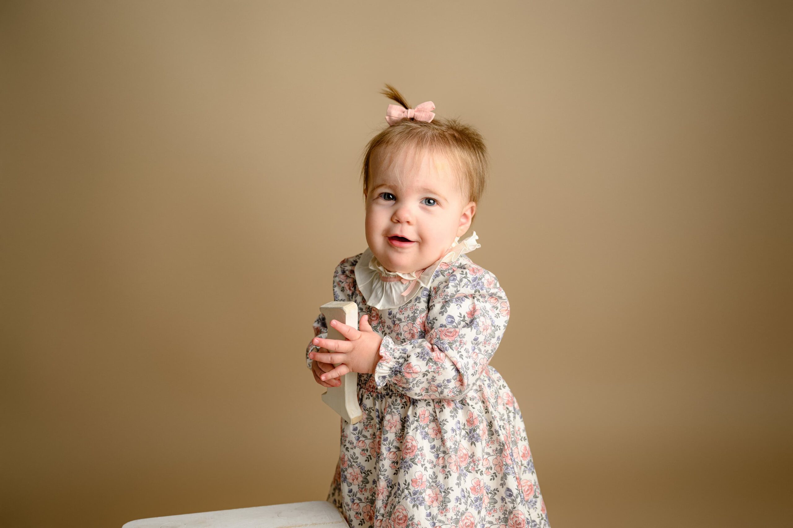 Molly wearing a flower dress holding a number one prop during her milestone session with a little pink bow in her hair