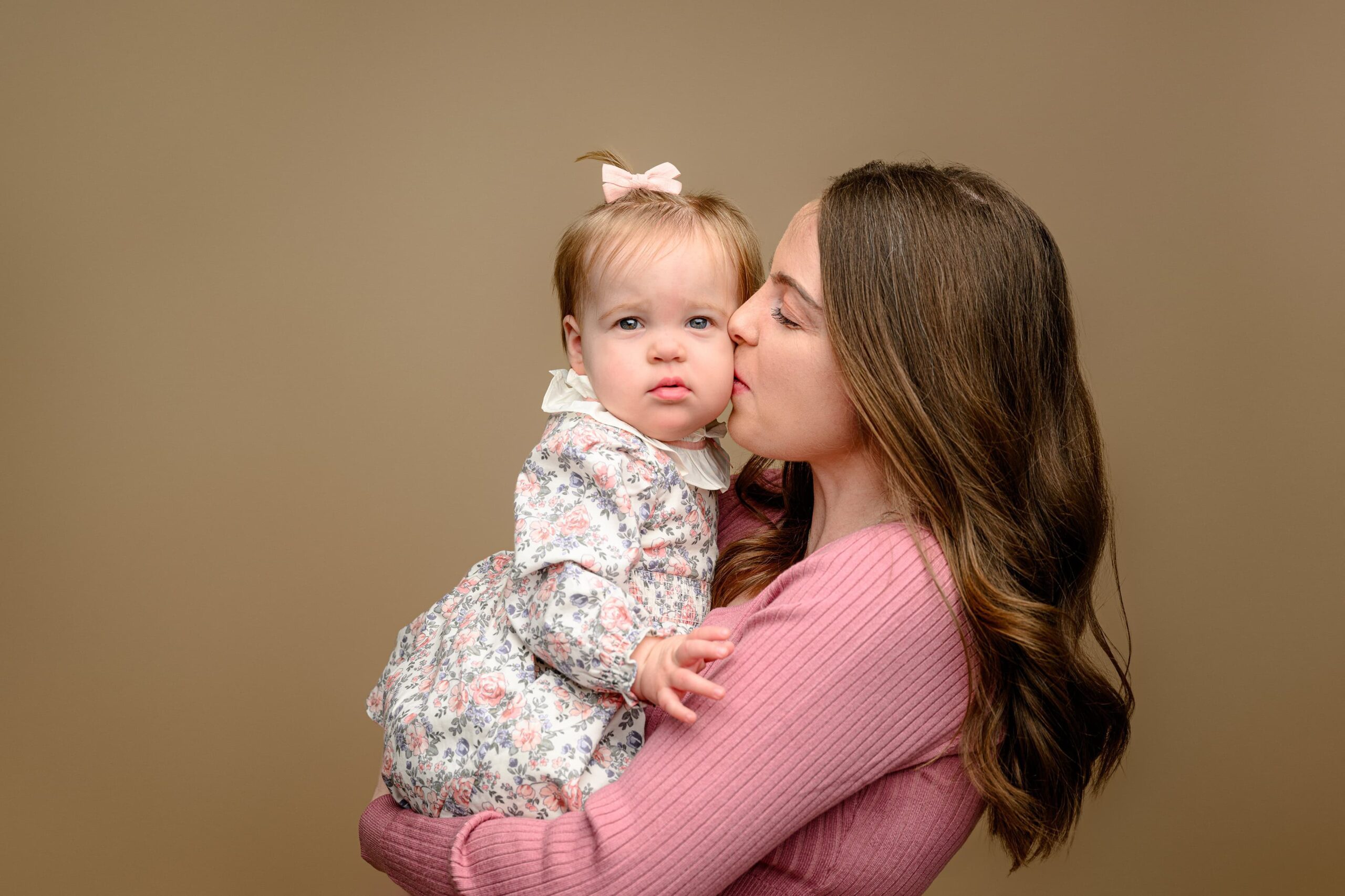 mom with long dark hair wearing a pink dress kisses her daughter on the cheek during their family mini photo session