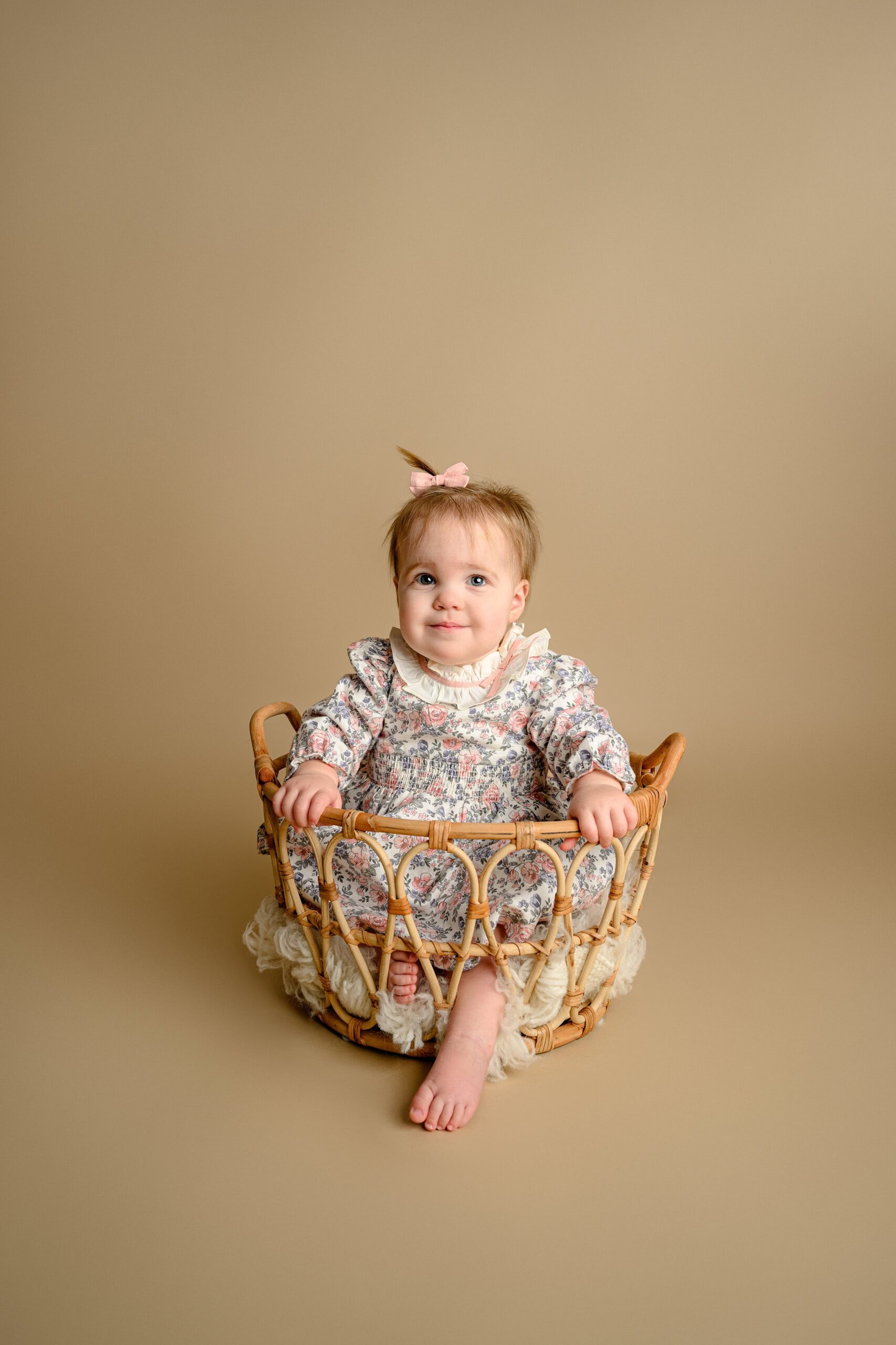 smirking as she plays in the basket a little girl sits with one foot out poking out during her photo session wearing a flower dress with a larger collar and a pink bow in her hair