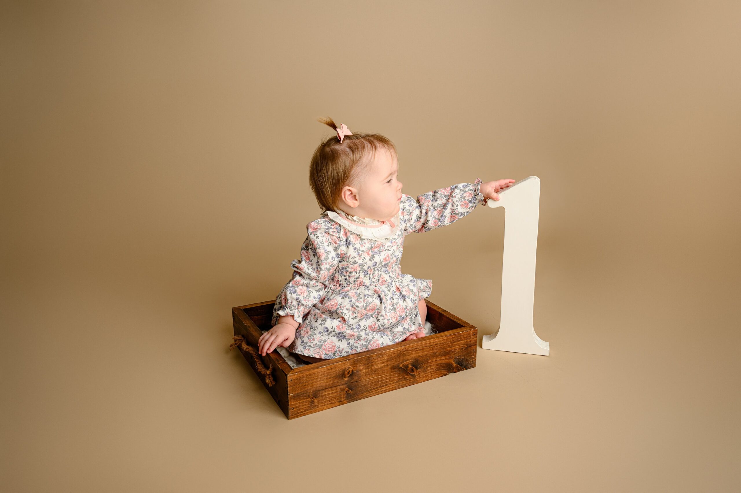 little baby girl wearing a flower dress sitting in wooden box touching a large number one prop