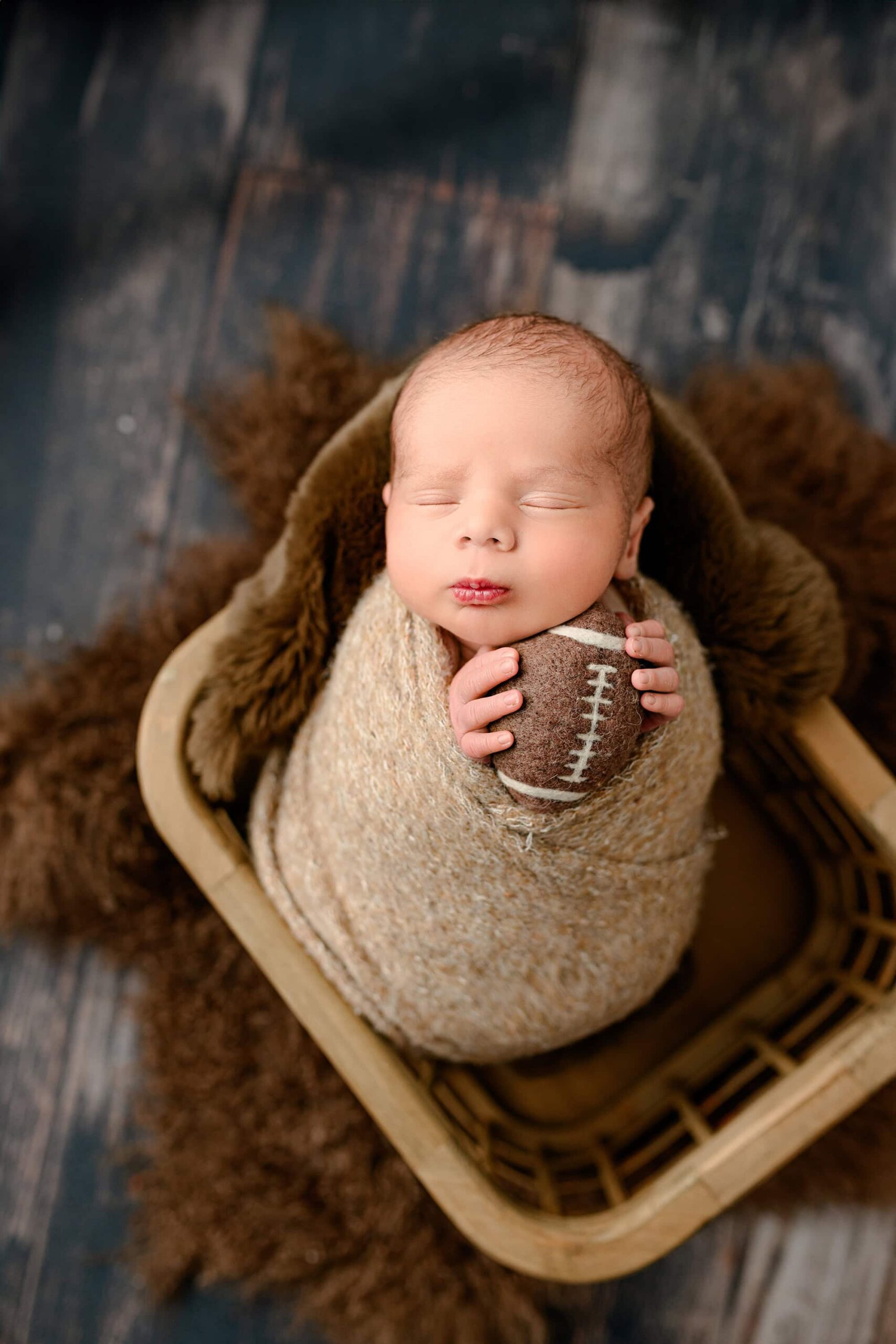 Newborn boy is wrapped in a brown wrap and placed in a basket filled with brown fur and on a wooden floor. he is holding a felted football close to his face.
