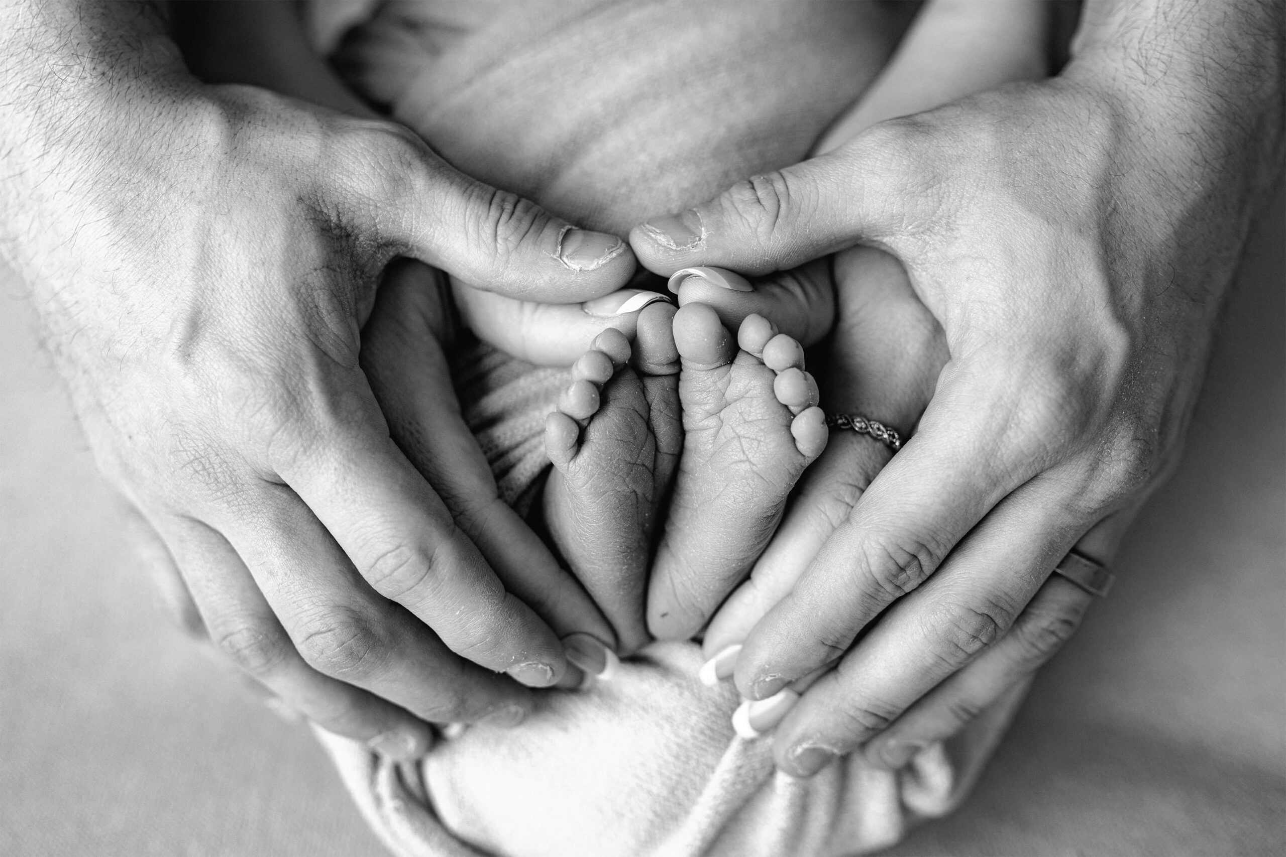 Black and white studio baby image of parents hands overlapping as they make a heart around their infant sons feet.