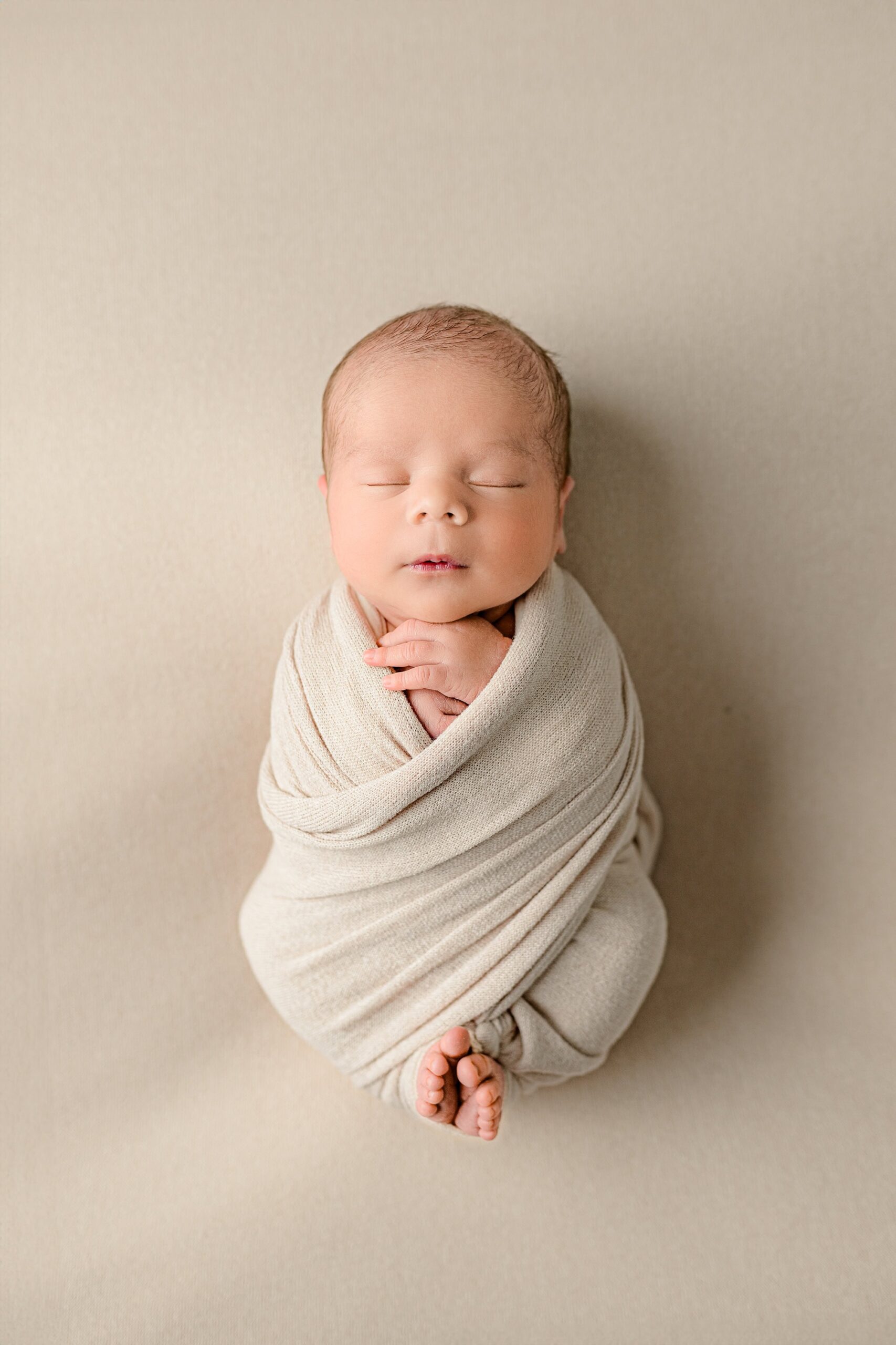 A newborn boy, wrapped in a cream-colored wrap with his toes peeking out, is lying on his back. The gentle pose showcases the baby's peaceful expression in this newborn session at a Waxhaw studio, with a soft cream background complementing the natural tones.