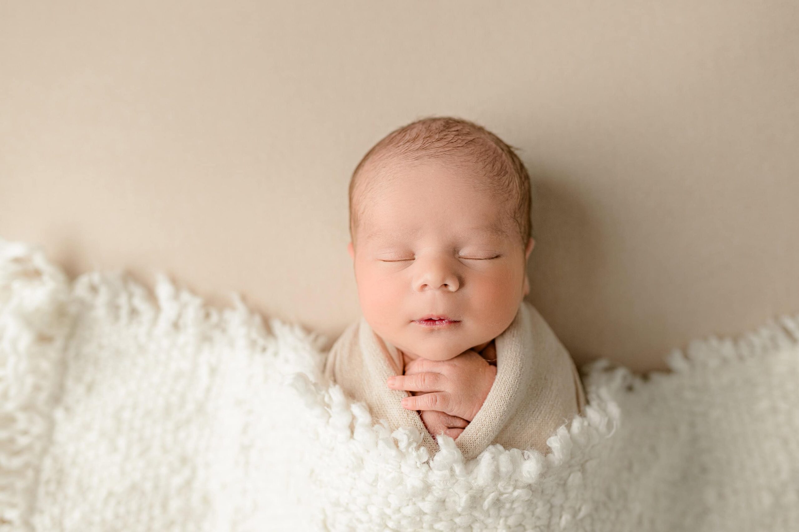 In this newborn session at a Waxhaw studio, a baby boy is wrapped in a cream-colored wrap and lying on his back with a cream blanket draped over him. The shot is taken overhead, highlighting the soft, serene setting of the baby against a matching cream background.