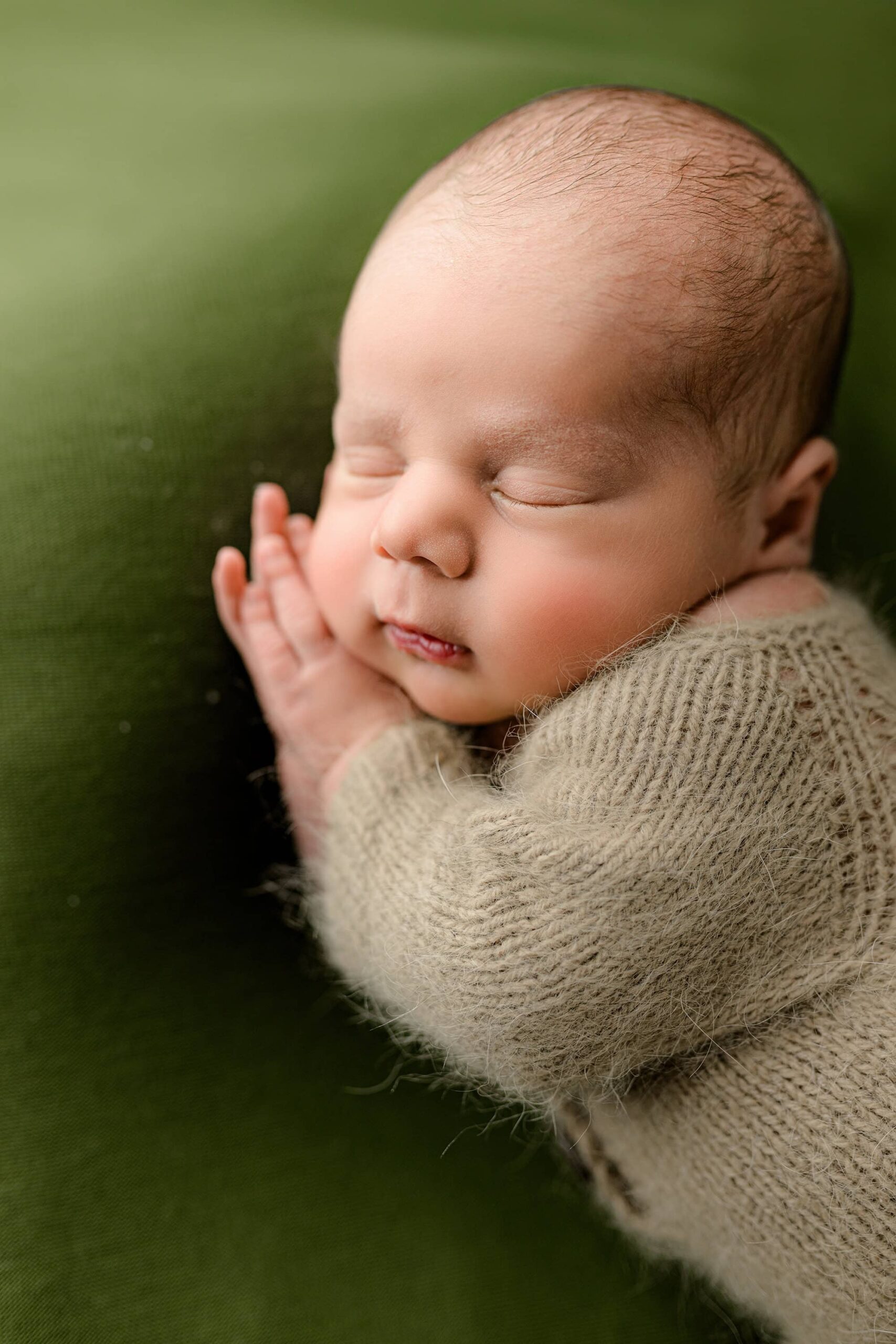 A close-up side profile of a baby boy in a brown knit footed romper in a Waxhaw studio. He is resting peacefully with his cheek on his hand, showcasing his sweet, serene expression against the soft green background.
