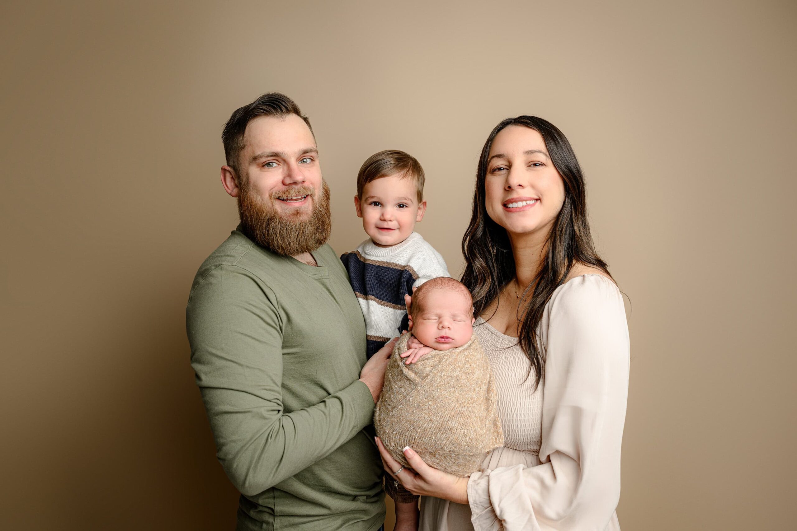 A family of four smiles at the camera. Dad holds their older son, who is wearing a striped sweater, while mom, dressed in a light brown dress, holds their newborn baby. The family is posed in front of a brown background, capturing a joyful, loving newborn session moment together.