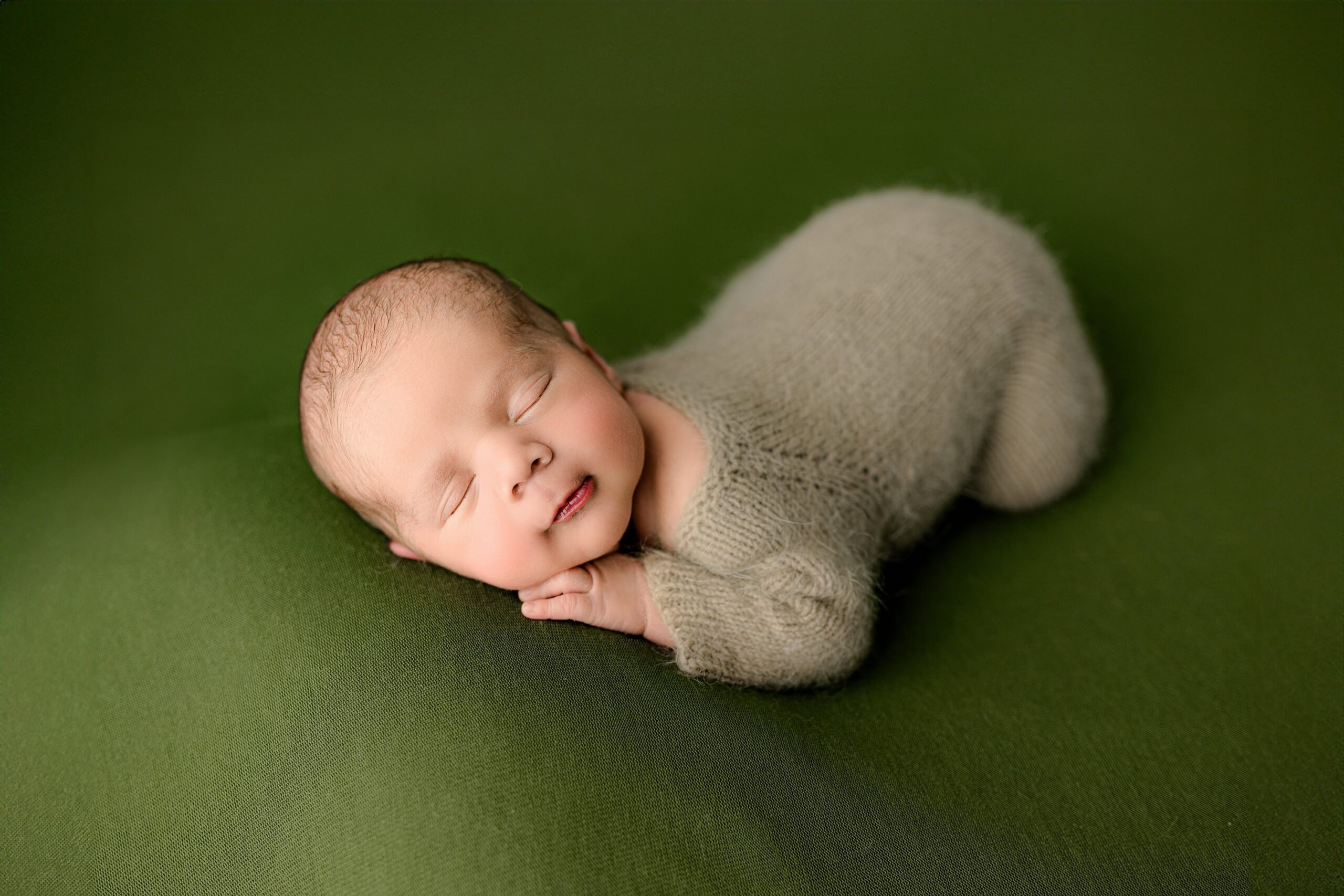 A baby boy dressed in a brown knit footed romper, lying on his belly. His hand is resting under his cheek, and his bottom is raised slightly in the air as he snuggles into the cozy pose on a green backdrop.