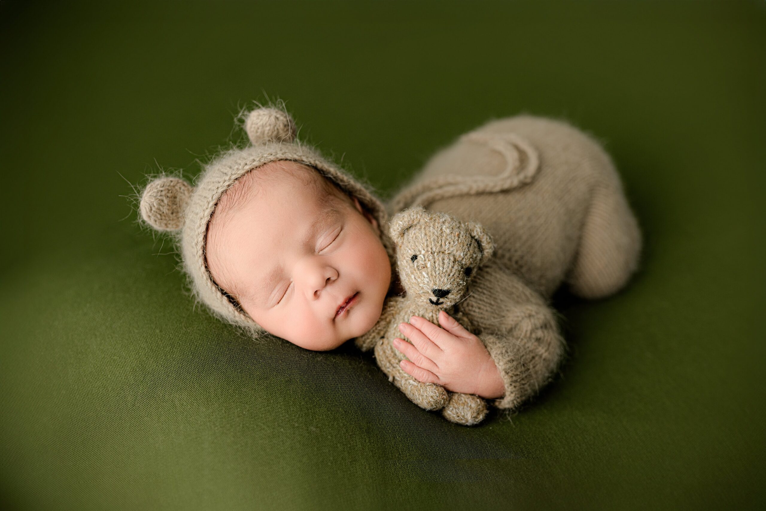 A baby boy dressed in a brown knit footed romper, with a matching brown hat featuring teddy bear ears. He is lying on his belly on a soft green background, gently cuddling a stuffed bear. His left arm is draped over the bear as he snuggles it in a peaceful pose.