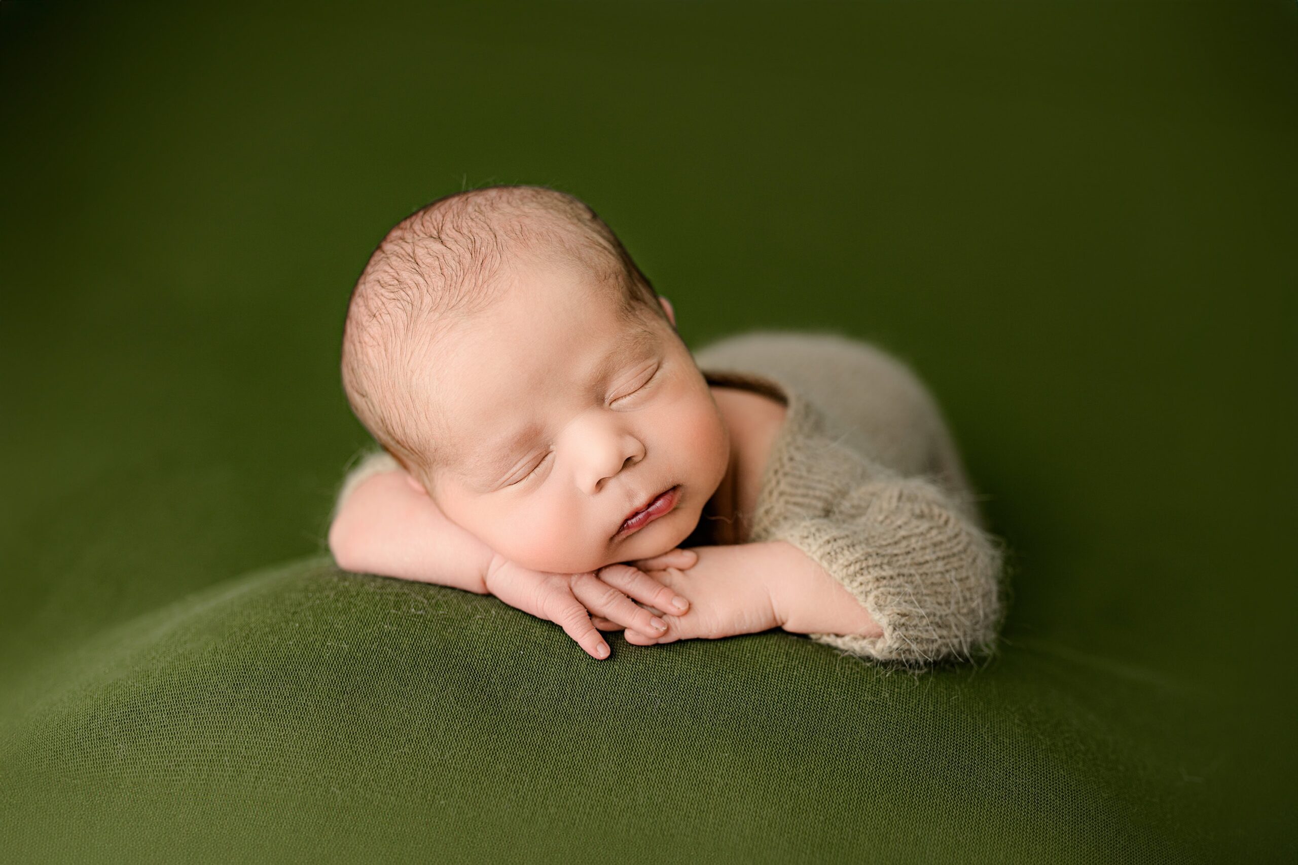 A baby boy in a brown knit footed romper, lying on his belly during his newborn session. His head is resting on his hands, which are placed under his chin, creating a sweet and relaxed pose on a green background.