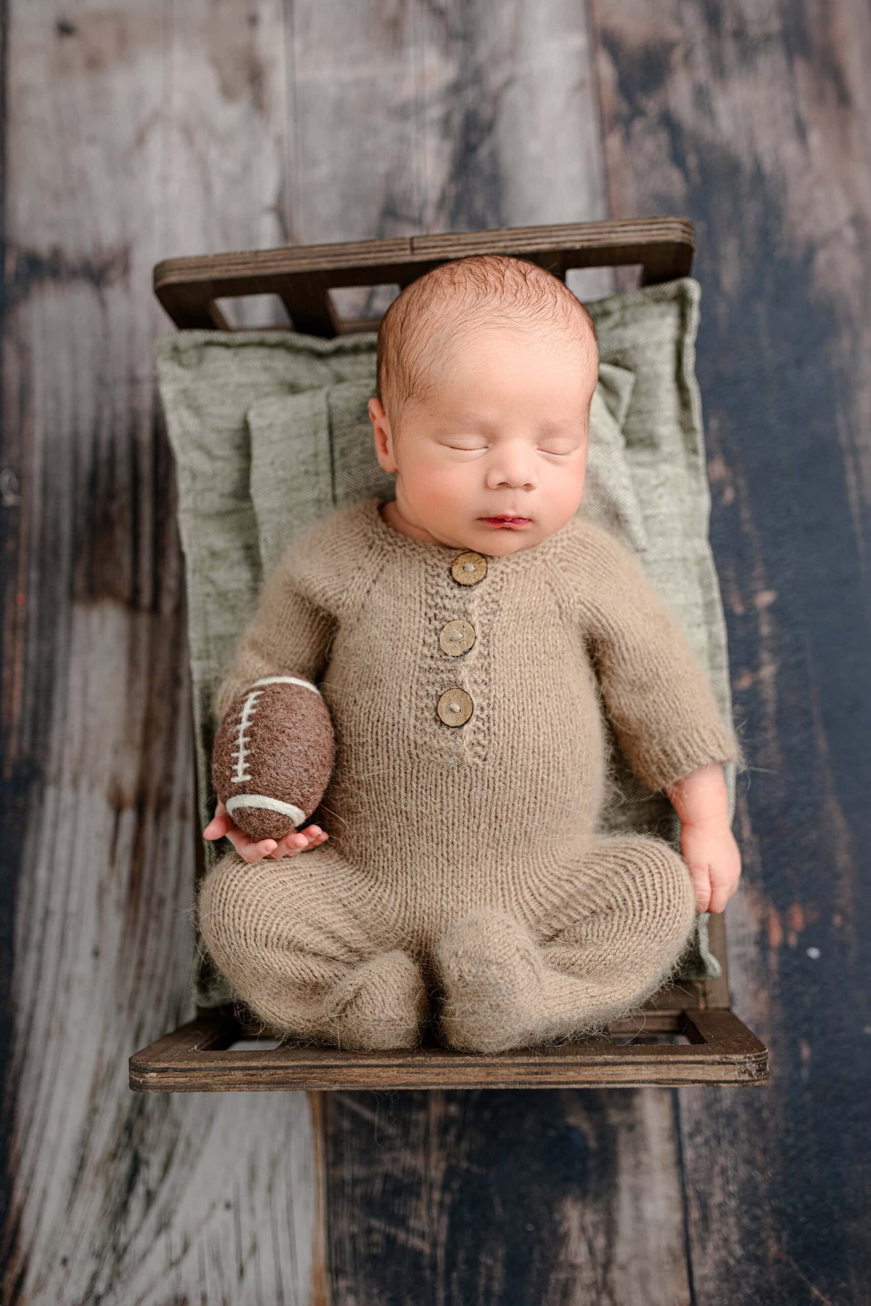 In this newborn session, a baby boy dressed in a brown knit footed romper is lying on a brown bed with green bedding on a wood floor. He is holding a small felted football, and the full image captures the peaceful scene of the baby and bed, showcasing the cozy atmosphere of the session.
