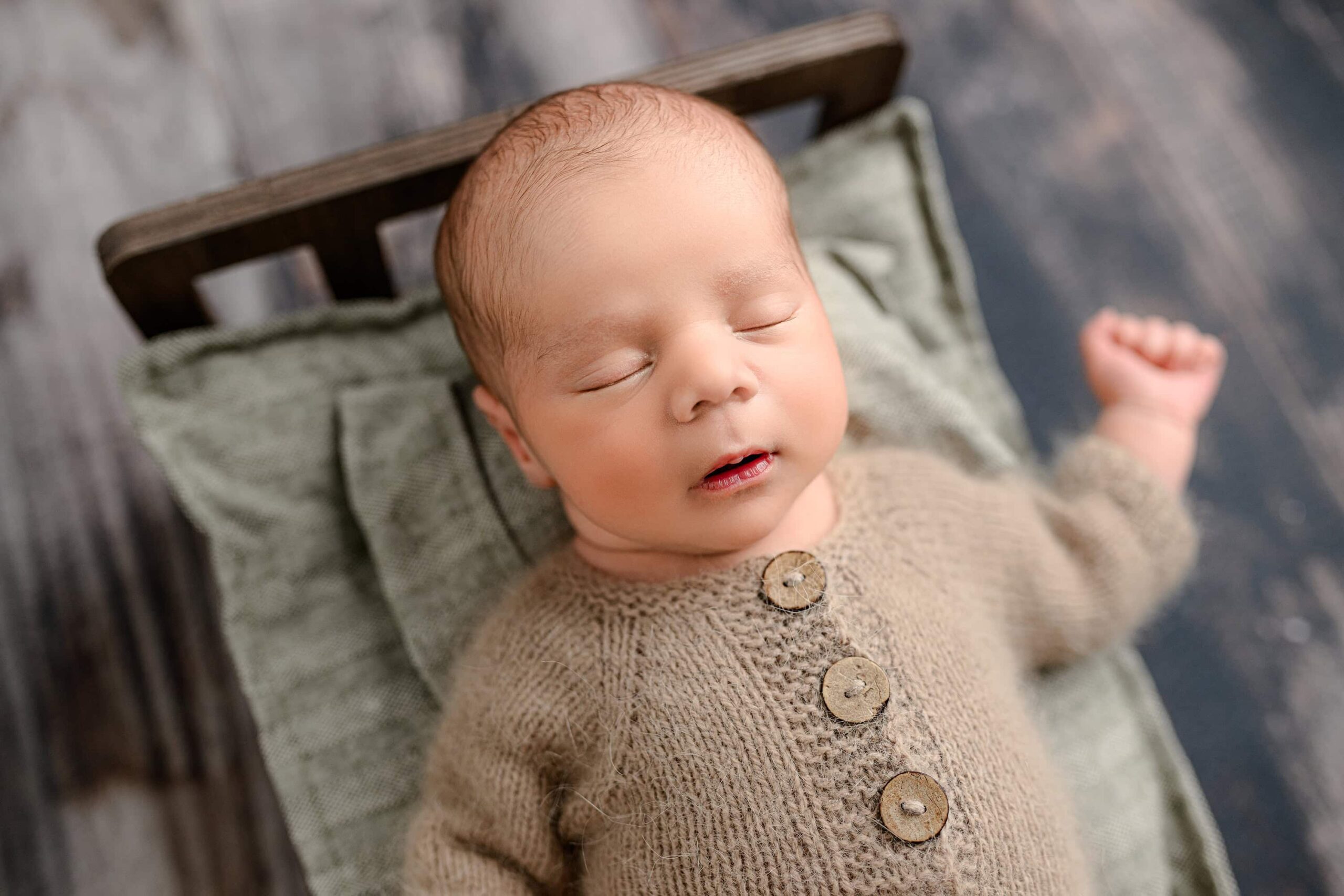 A half-body shot from a newborn session at a Waxhaw studio, featuring a baby boy in a brown knit footed romper. He is lying on the brown bed with green bedding, with his arm extended out to the side in a relaxed pose.