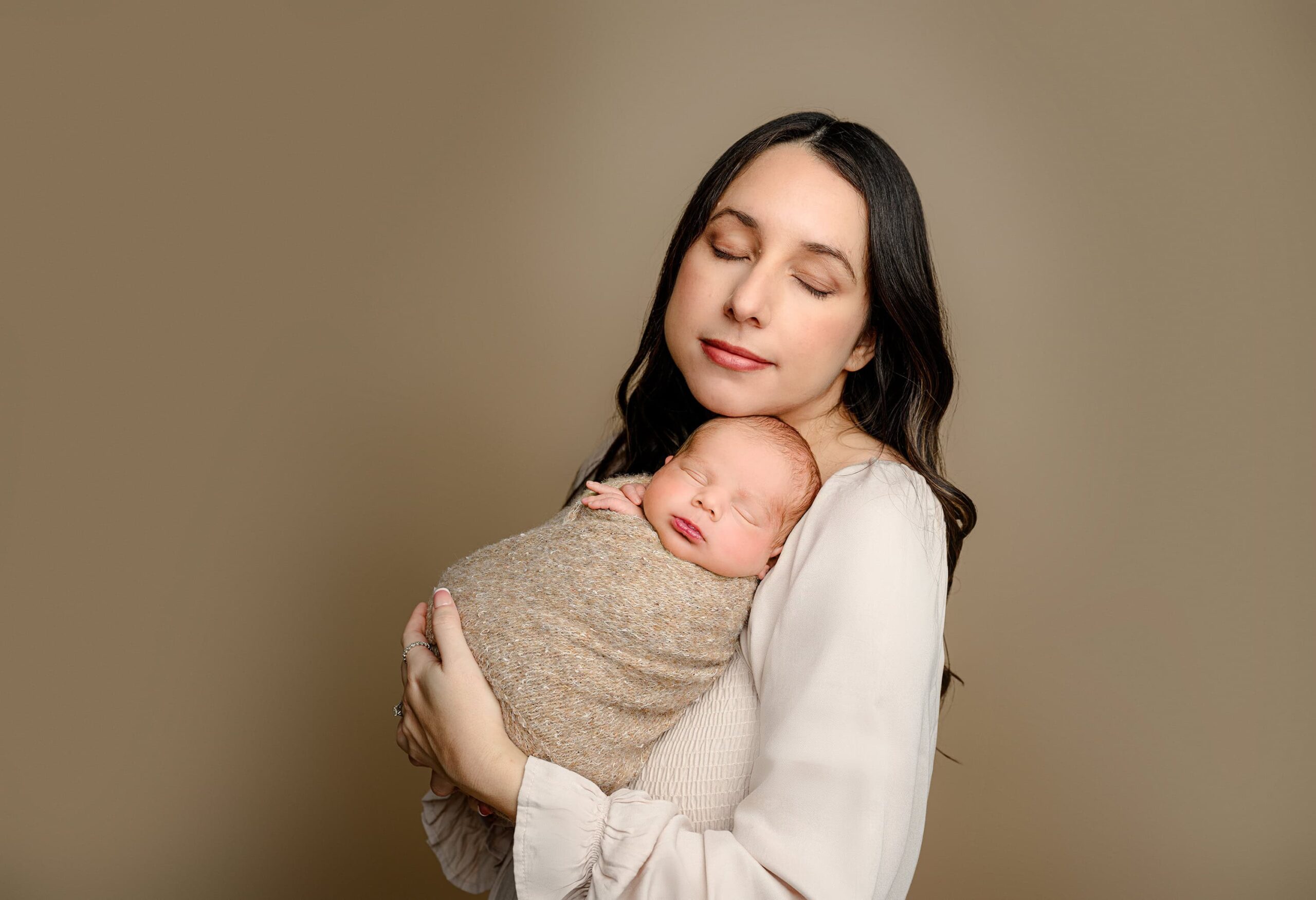 Mom, wearing a light brown dress, holds her newborn baby boy against her shoulder. Her chin is gently resting close to his head, and both mom and baby have their eyes closed, creating a serene and tender moment of connection between them.