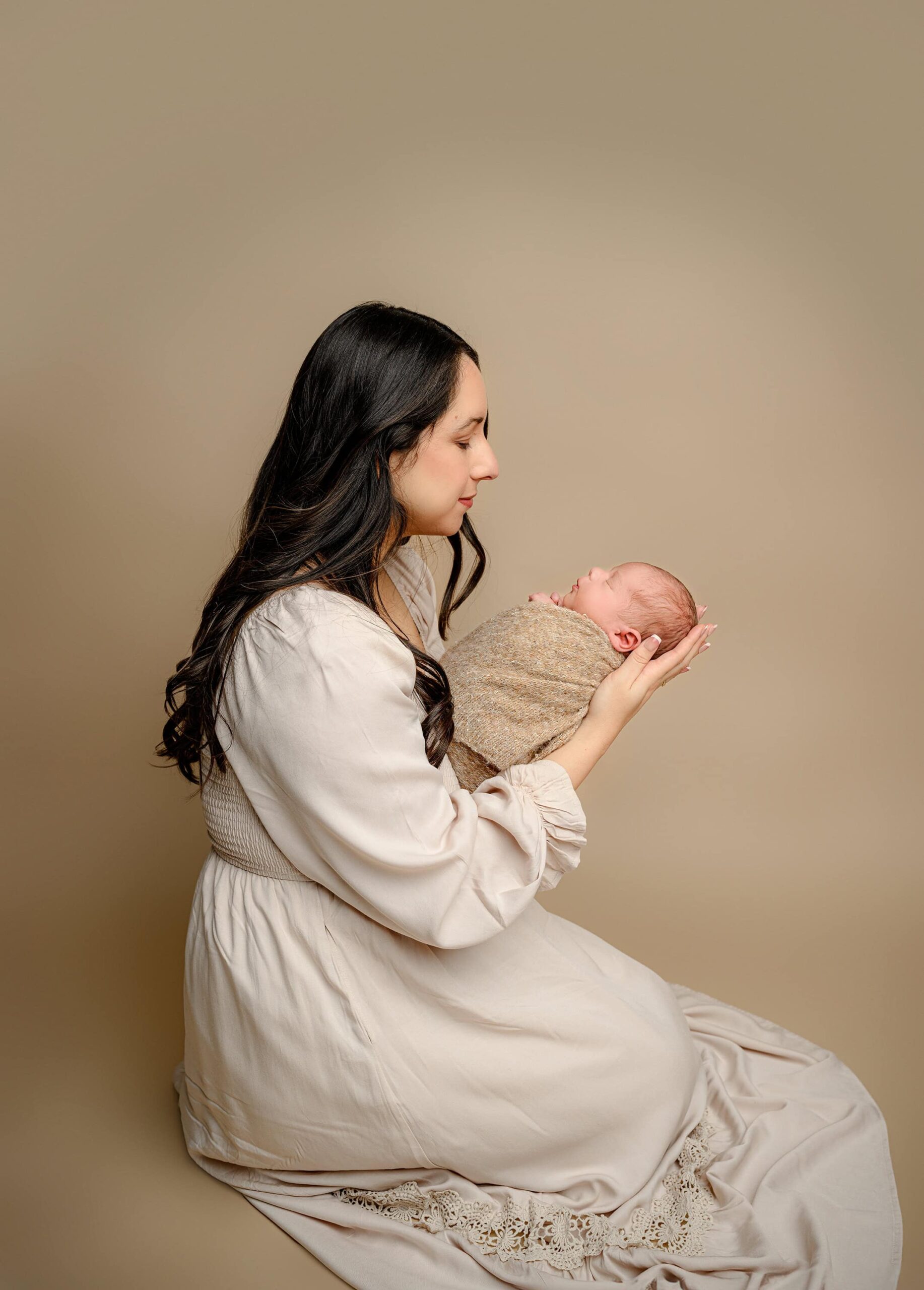 A Waxhaw, NC studio full body profile shot of mom kneeling and holding her newborn son in front of her, looking down at him. The baby is wrapped in a brown wrap, and mom’s expression shows pure affection as she adores her little one.